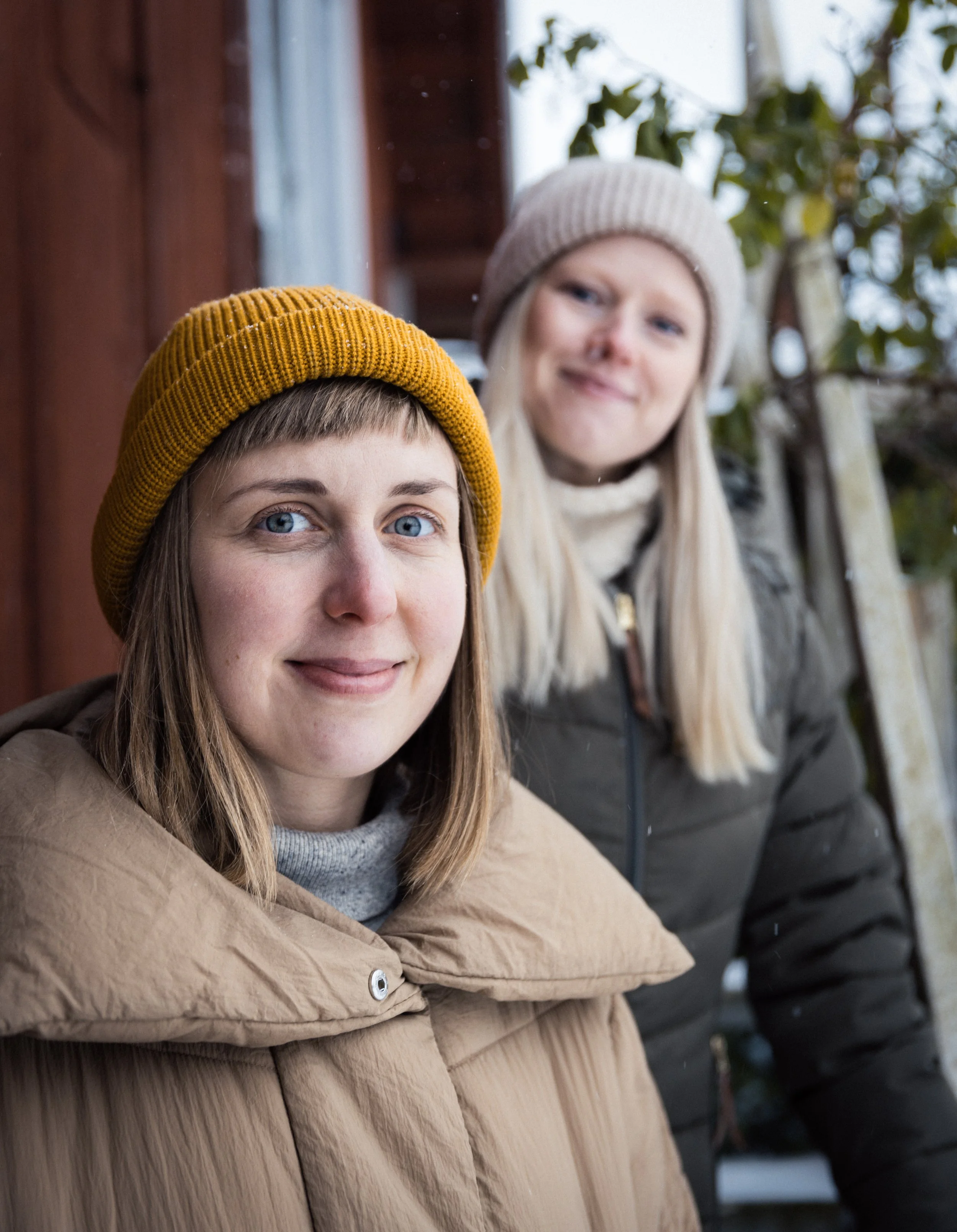 Two women outdoors in winter clothing, one wearing a mustard-yellow knit beanie and beige jacket, the other in a light gray beanie and dark gray puffer coat, standing near a wooden fence with greenery in the background.