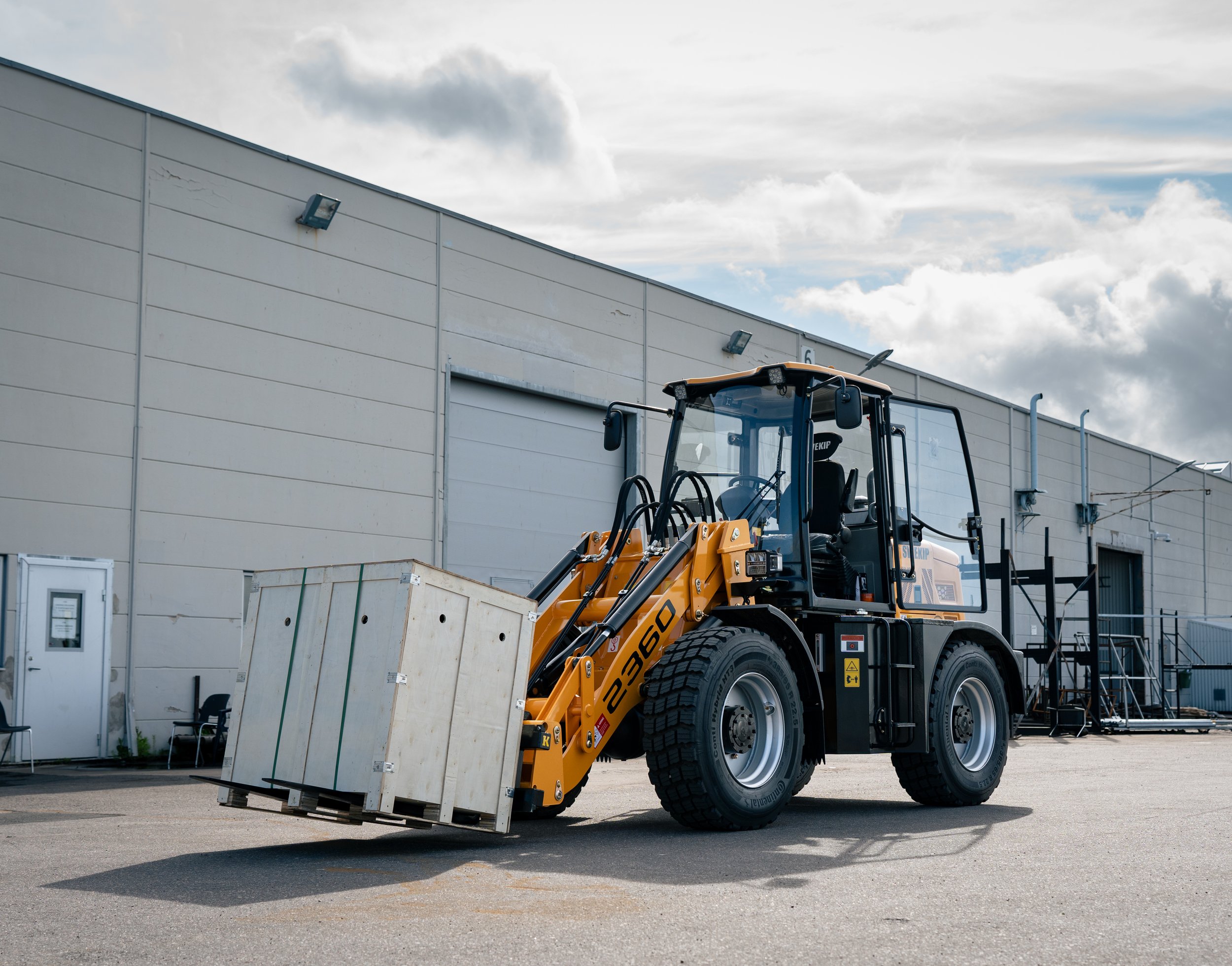 A yellow compact wheel loader carrying a large white crate outside a warehouse on a cloudy day.