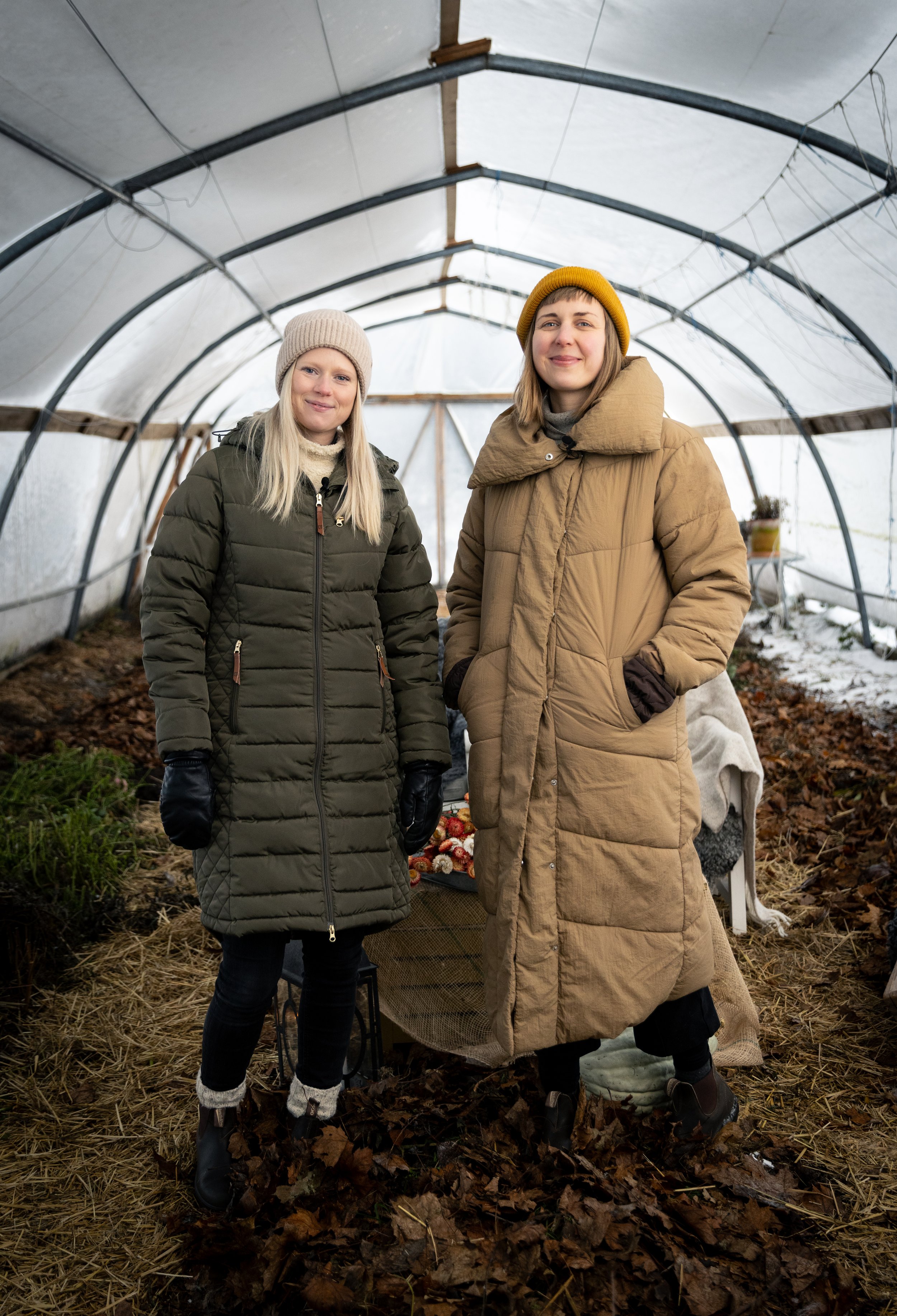 Two women standing inside a greenhouse, dressed in winter coats and hats, with plants and gardening supplies around them.