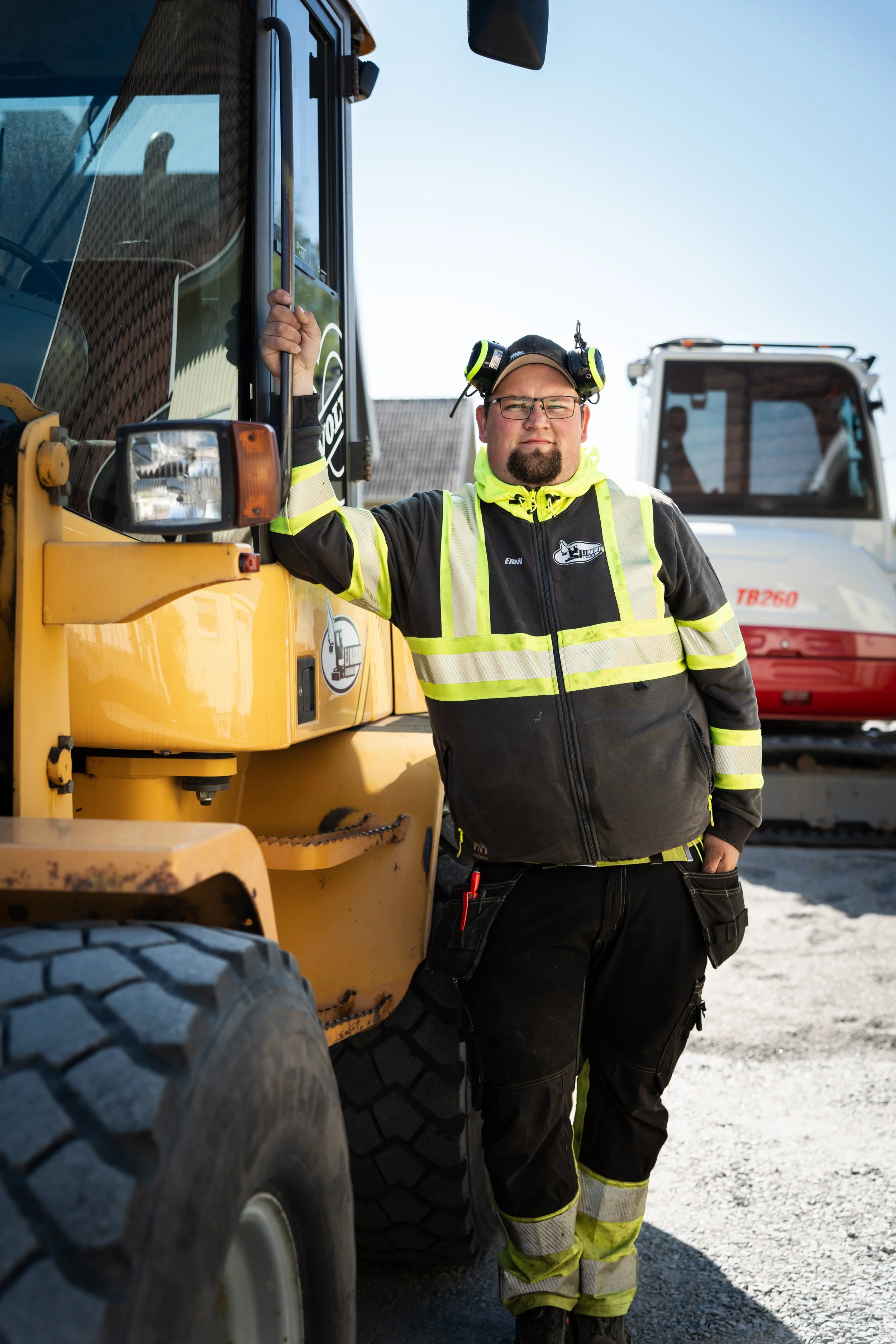 A construction worker wearing a black and yellow jacket, safety glasses, and a helmet with headphones, leaning against a yellow construction vehicle at a worksite with another vehicle in the background.