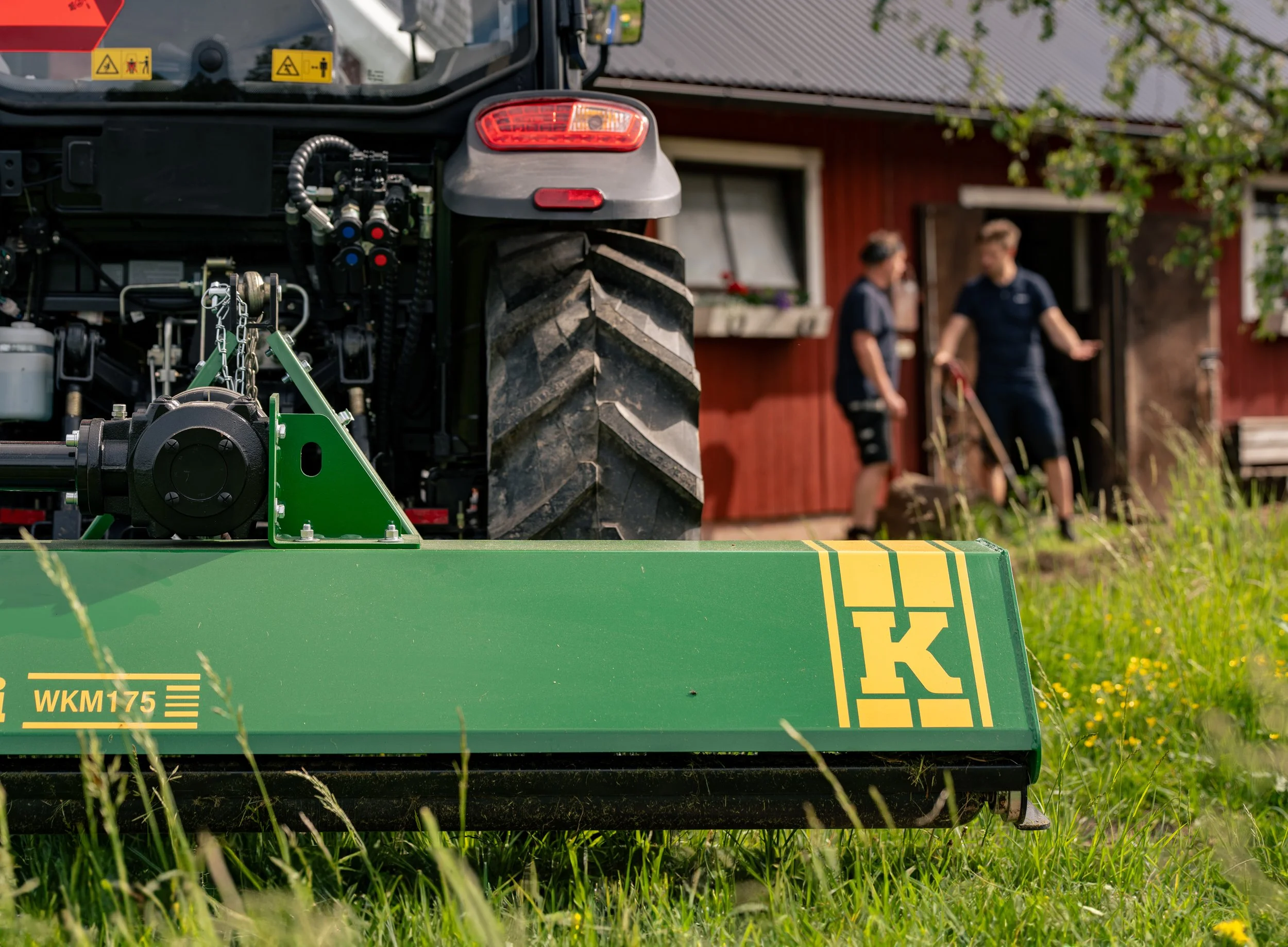 Close-up of a green agricultural machine, with two people in the background near a red wooden house.