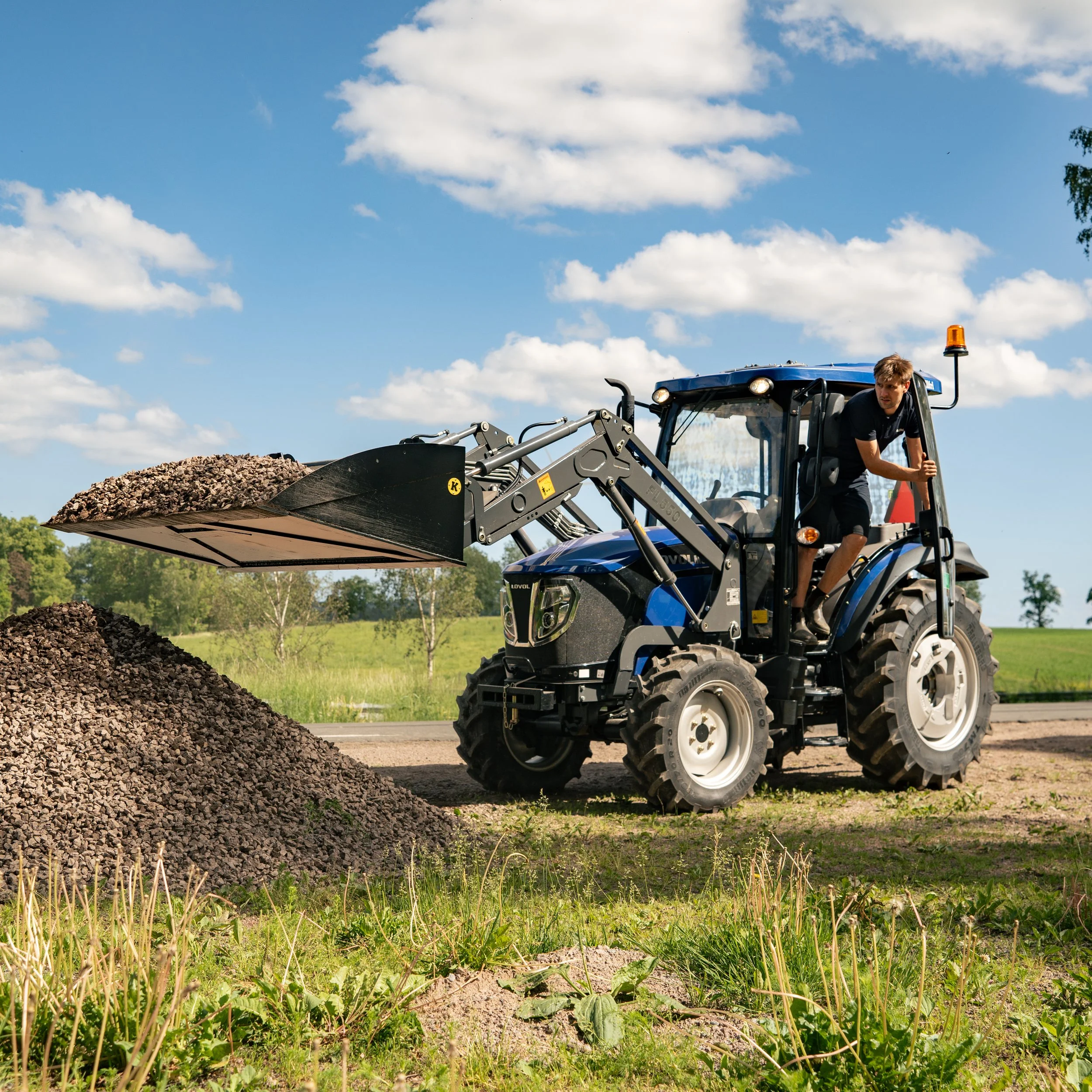 A man operating a blue compact tractor with a front loader, unloading gravel onto a pile in a grassy field on a sunny day with a blue sky and scattered clouds.