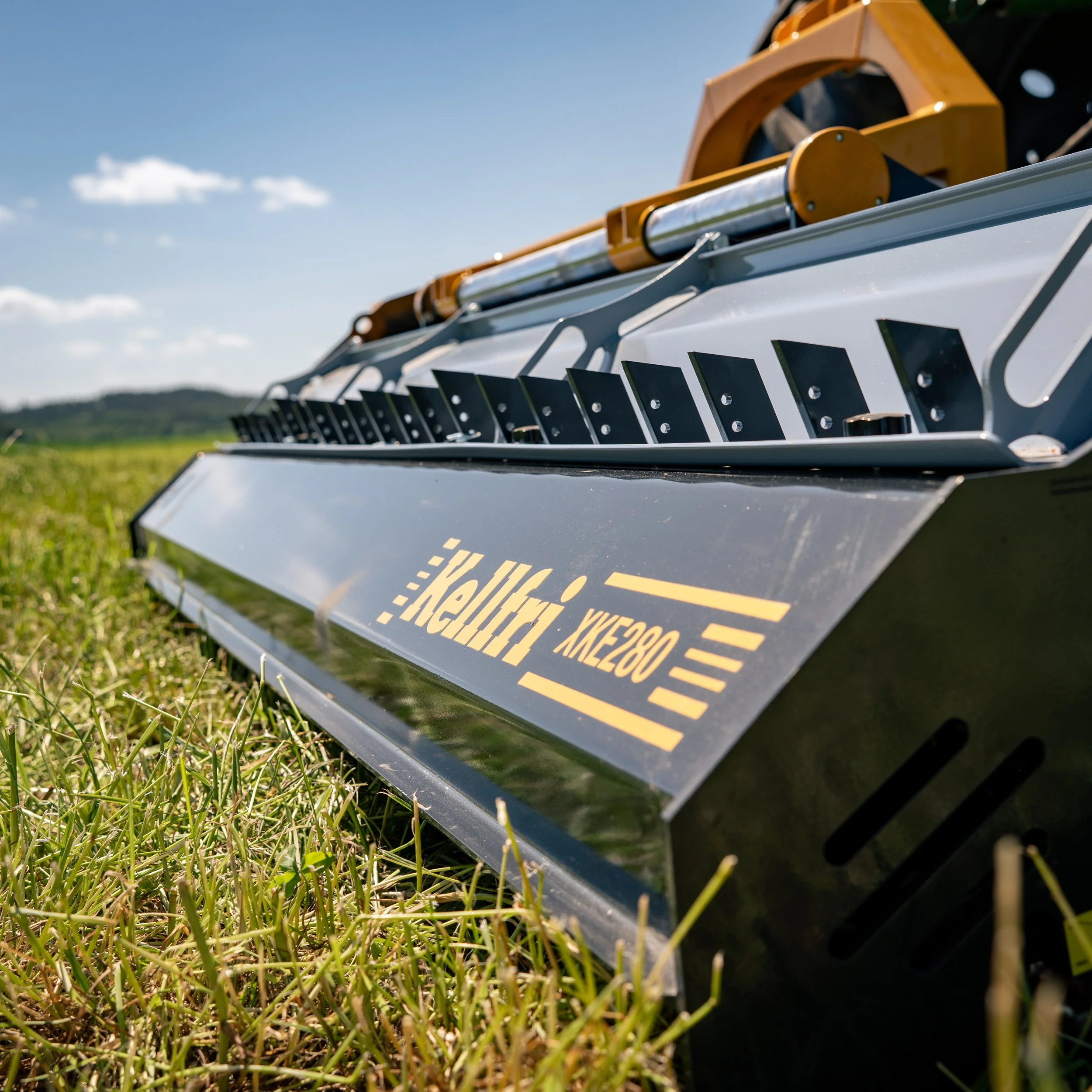 Close-up of a Hellcat lawn mower attachment on a grassy field with a clear blue sky and distant hills.