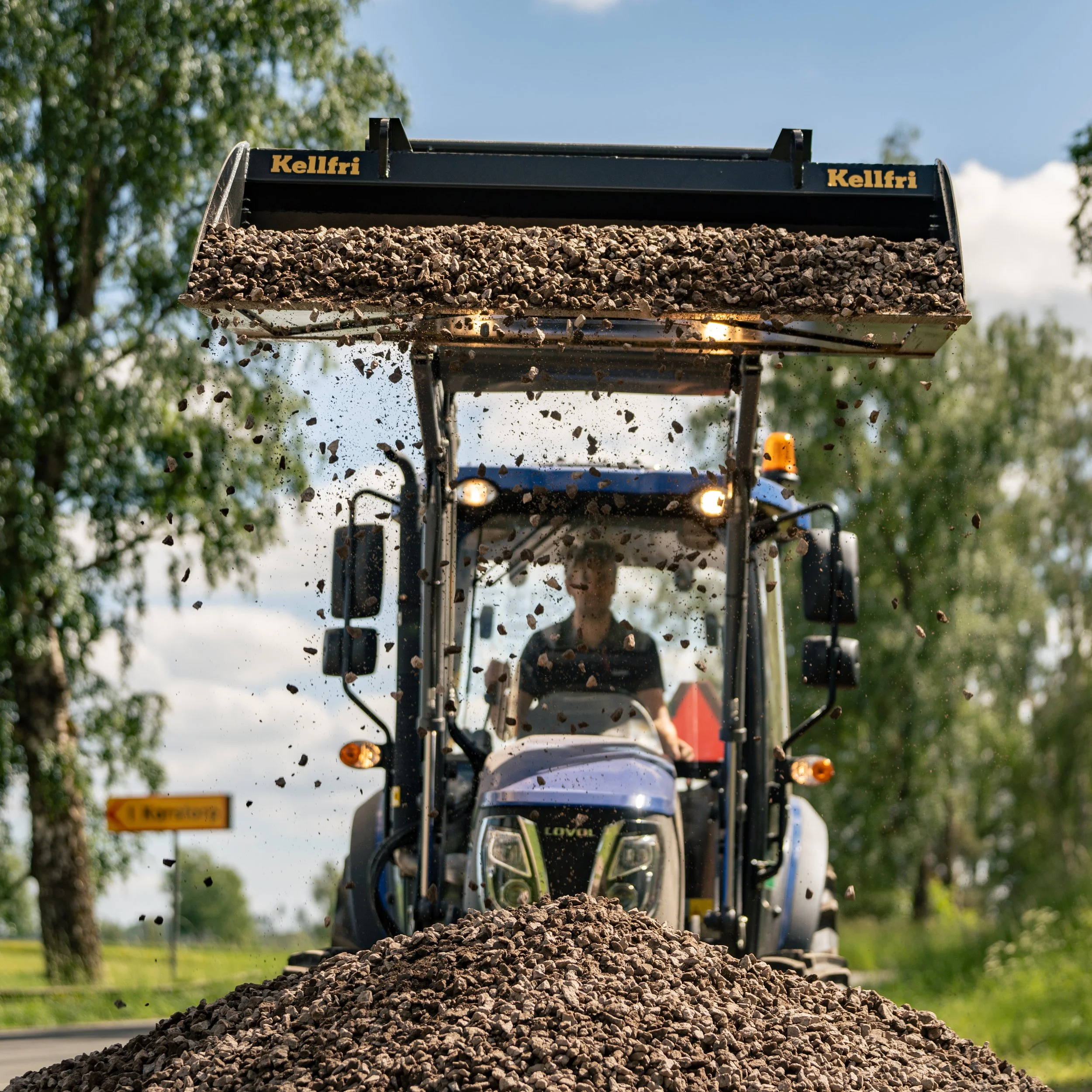 A tractor spreading gravel on a road, with a person driving in the cabin, surrounded by trees and a blue sky.