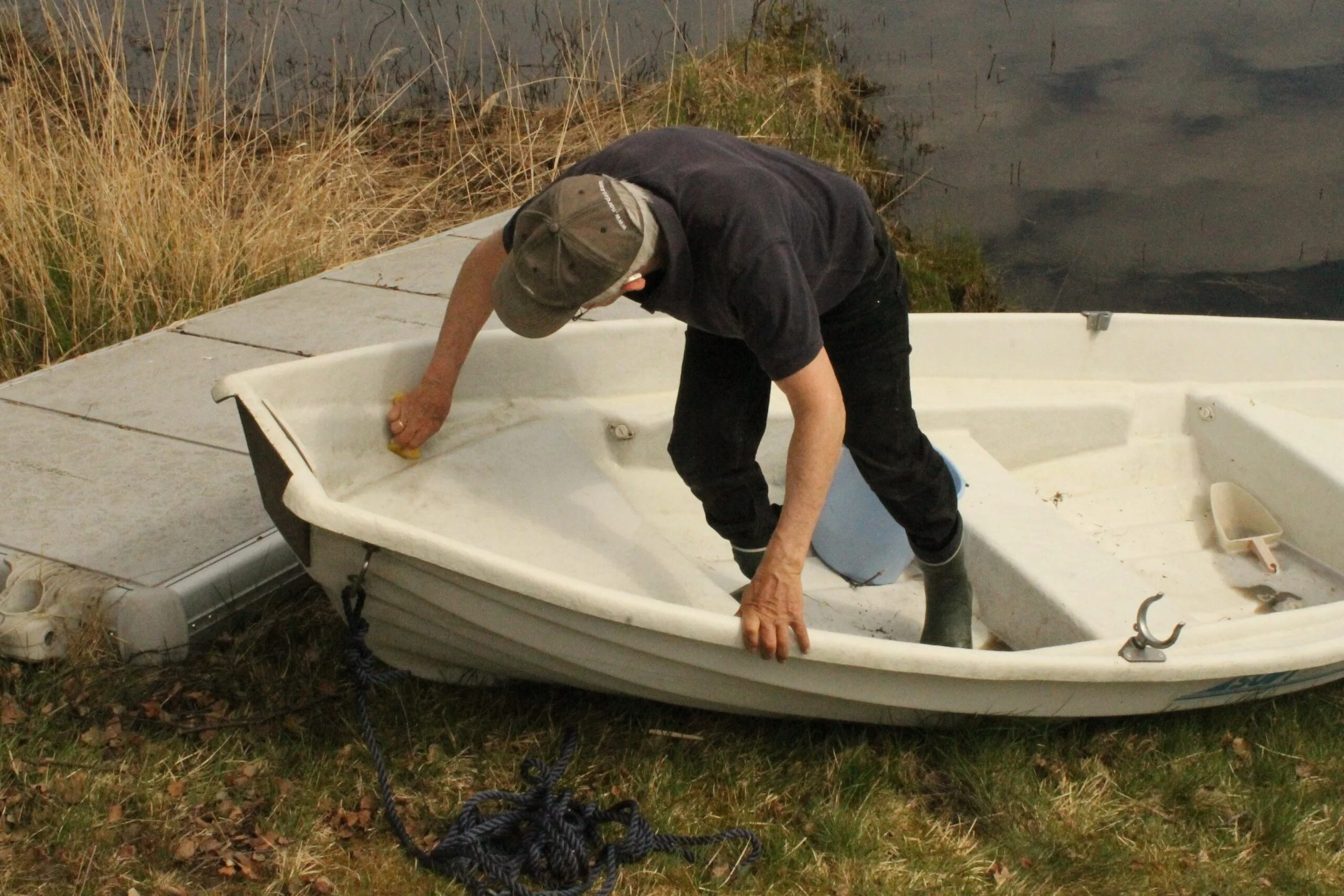 En mann vasker en liten hvit båt ved elvebredden, omgitt av gress og vann.