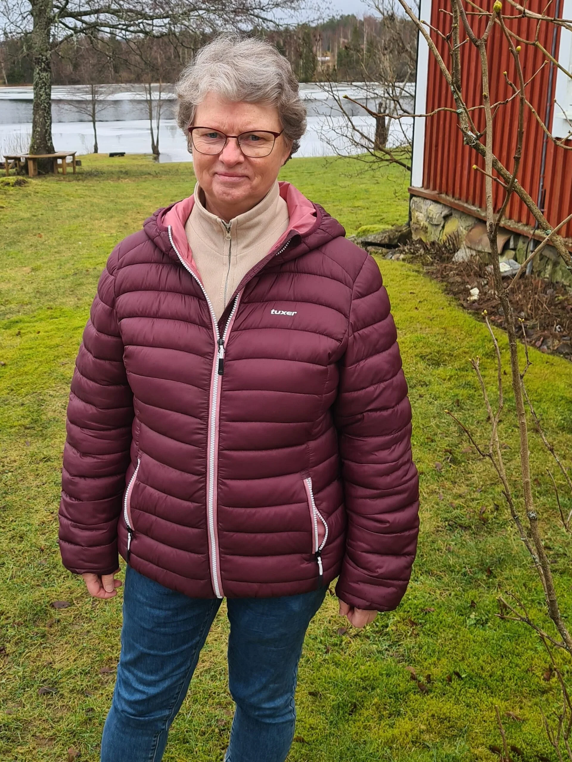 En kvinne i en burgunder vindjakke og blå jeans står utendørs på en grønn plen, med vann og trær i bakgrunnen.