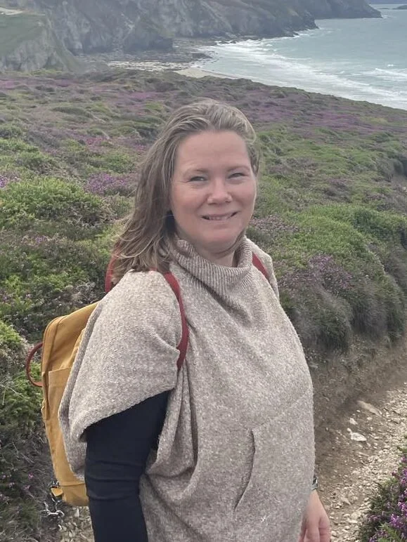 En kvinne som står på en kyststi med blomster på siden, utsikt over sjø og klipper i bakgrunnen.