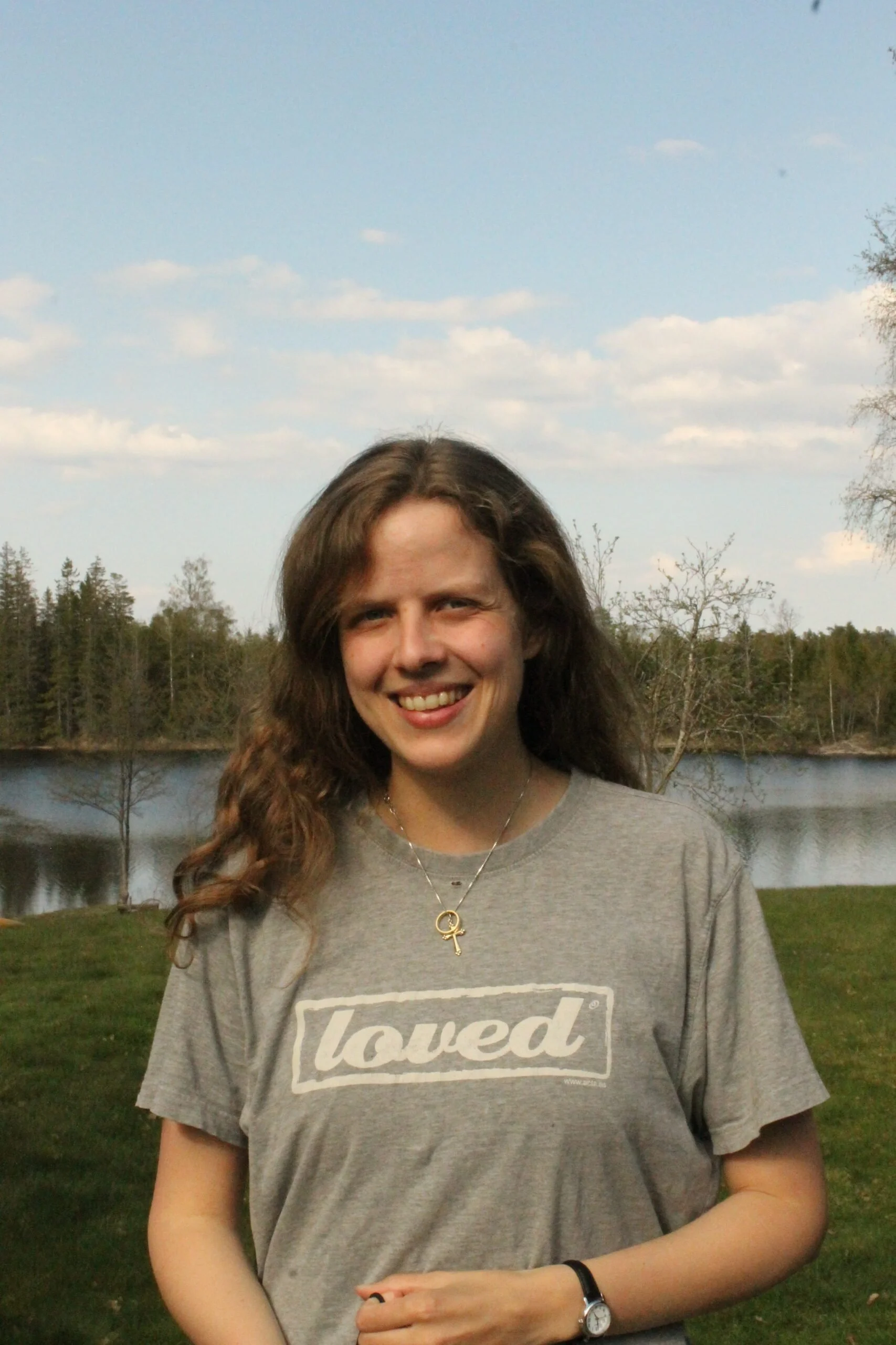 Et smilende ungdoms- eller unge voksen kvinne med langt, bølgete brunt hår, iført en grå t-skjorte med teksten "loved" og et hjerte og Venus-tegn kjede, står utendørs ved en innsjø med trær og en åpen himmel i bakgrunnen.