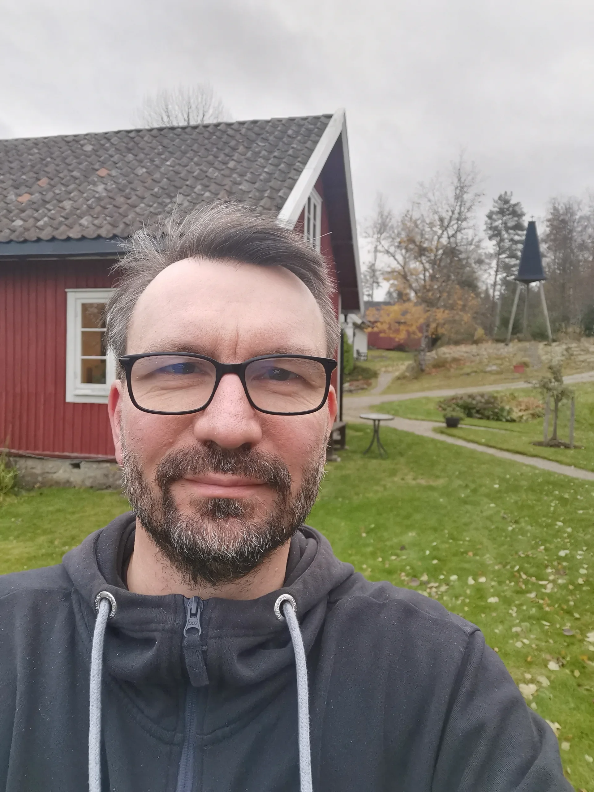 En mann tar et selfie foran et gammelt hus på en grå dag.