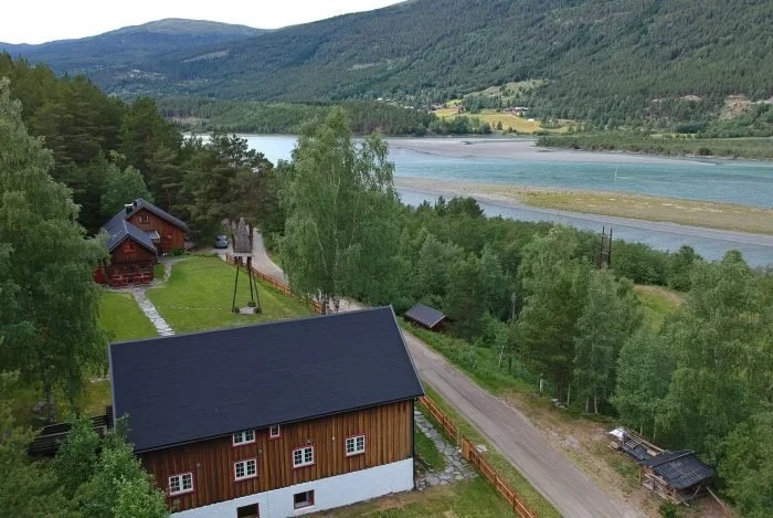 Utsikt over en gård med flere trehus omgitt av grønt landskap og en elv i bakgrunnen.