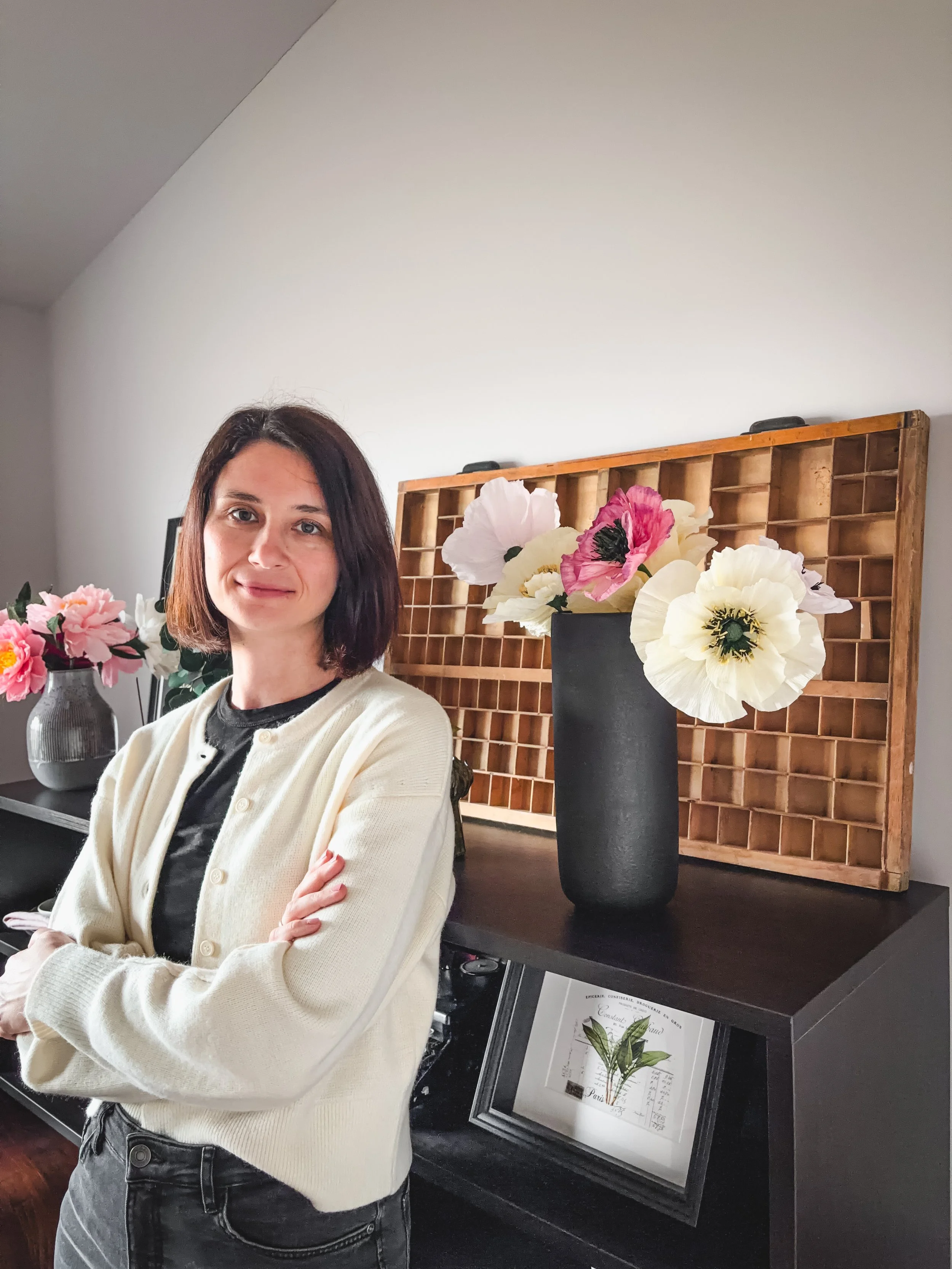 Eine Frau mit braunen, kinnlangen Haaren trägt eine schwarze Oberbekleidung und eine cremefarbene Strickjacke, steht in einem Raum mit weißen Wänden und einem schwarzen Regal, das dekorative Vasen mit pinken und weißen Blumen enthält, darunter eine große schwarze Vase im Hintergrund.