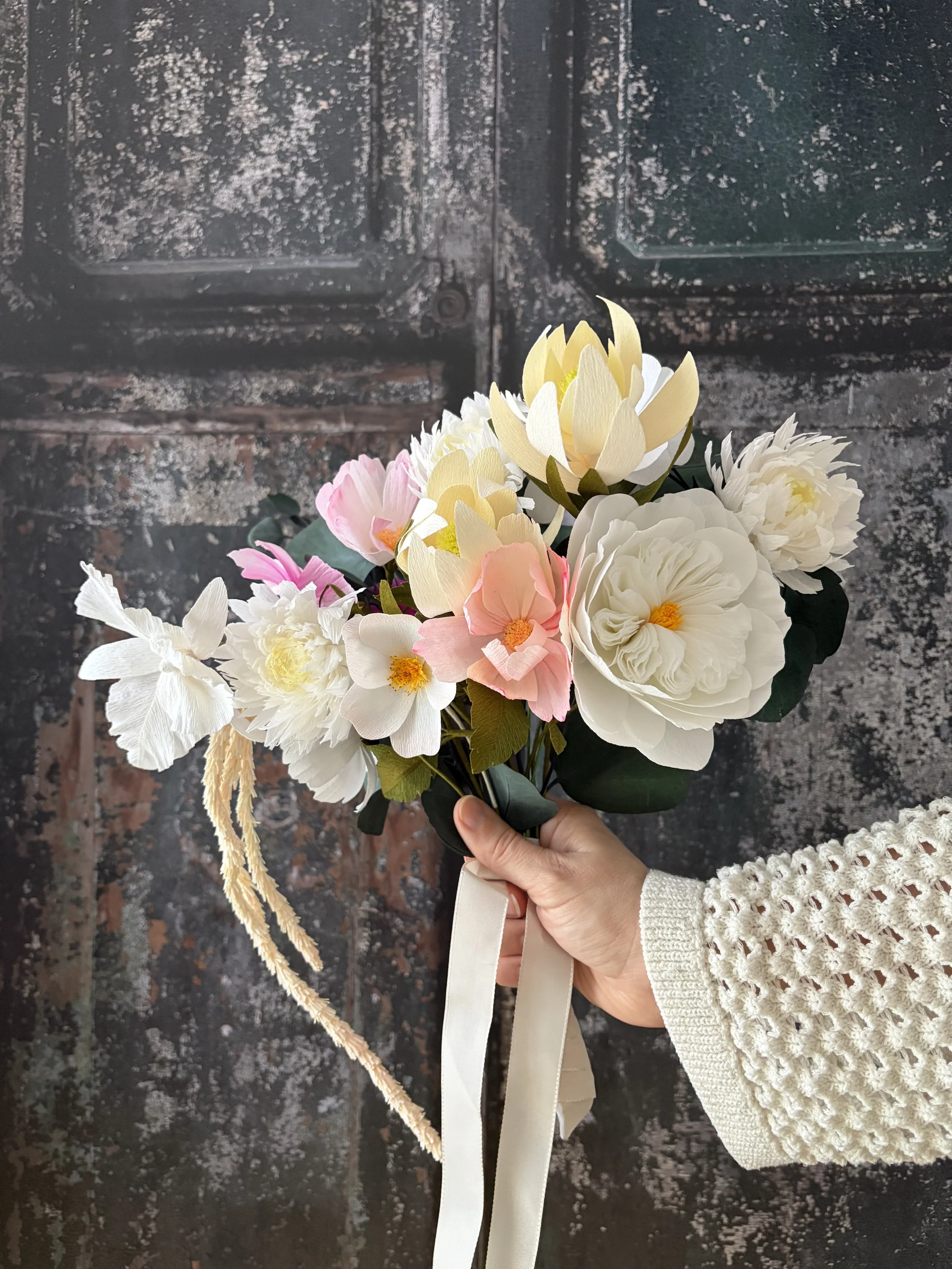 Eine Hand hält einen Strauß aus weißen, rosa und gelben Stoffblumen mit grünen Blättern vor einer dunklen, alten Tür.