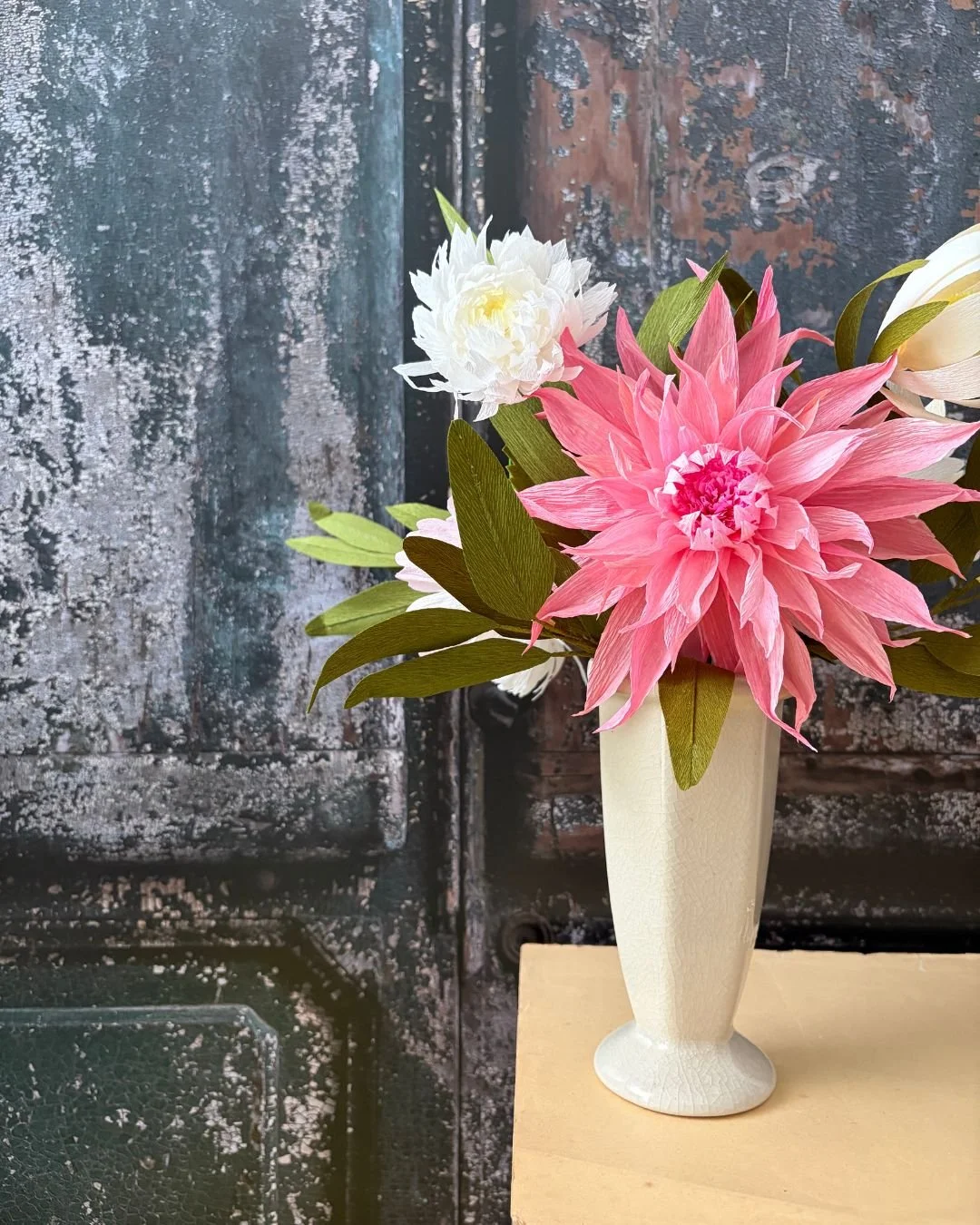 Blumenstrauß mit großen pinken, weißen und cremefarbenen Blüten in einer weißen Vase auf einem Holztisch gegen eine dunkle, alte Tür