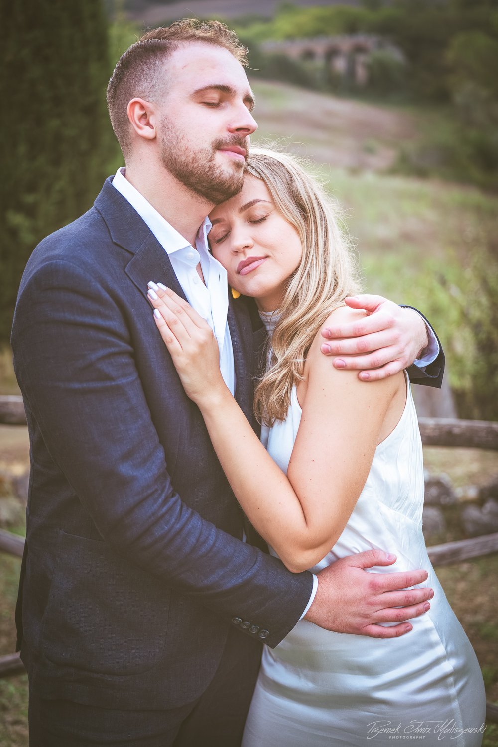 A man and woman embracing outdoors, eyes closed, with the man in a suit and the woman in a sleeveless dress, standing in front of a natural background with trees and a fence.