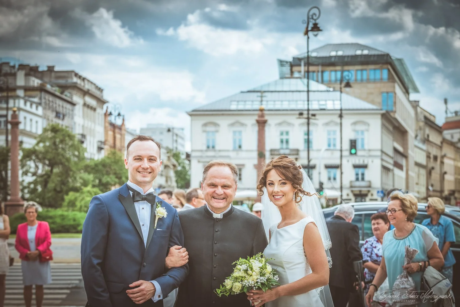 A wedding couple with a priest or officiant, standing outdoors in an urban area with onlookers in the background, city buildings, and a cloudy sky.