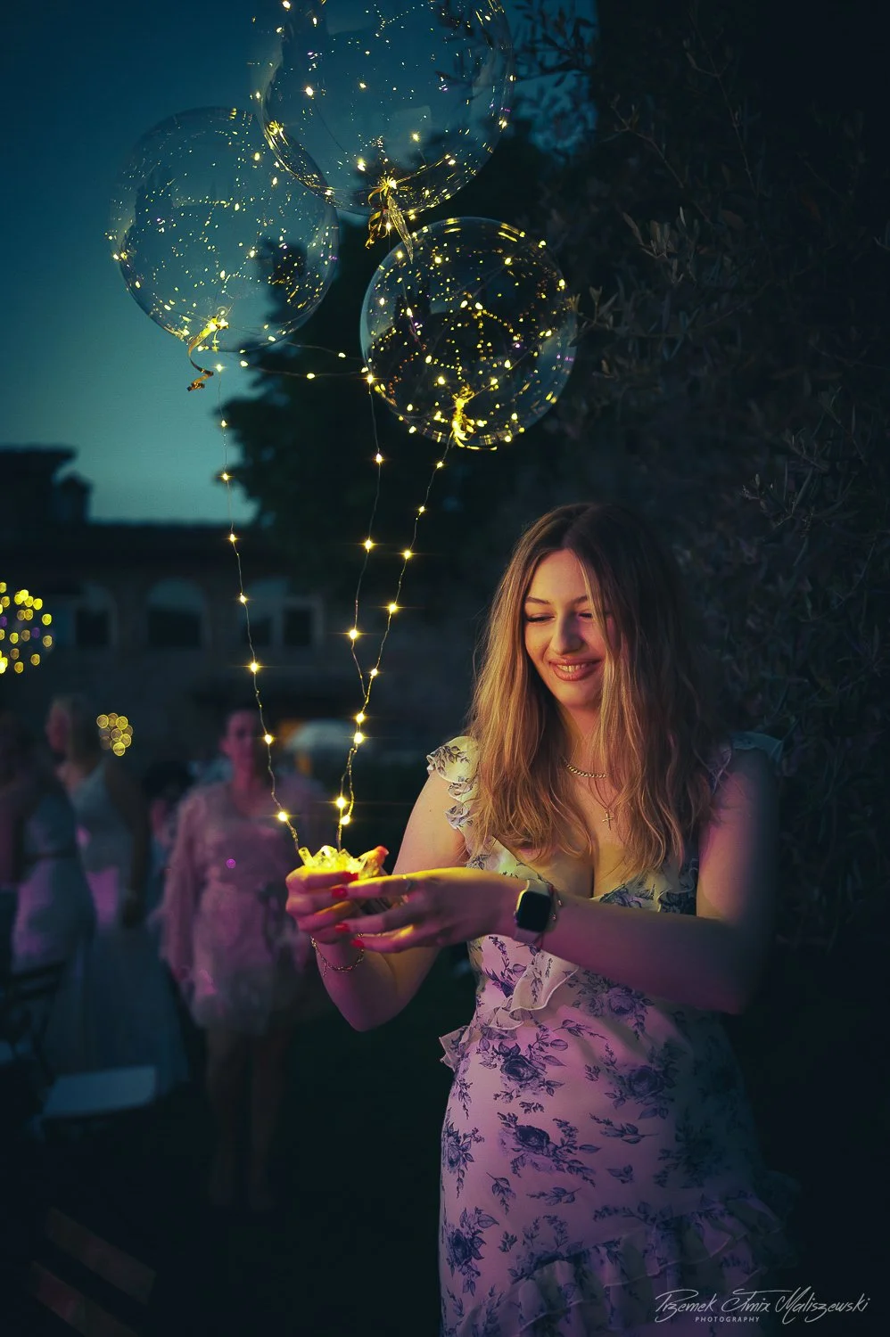 A young woman holding a tray with glowing string lights and balloons, smiling during an outdoor evening party with blurred guests in the background.