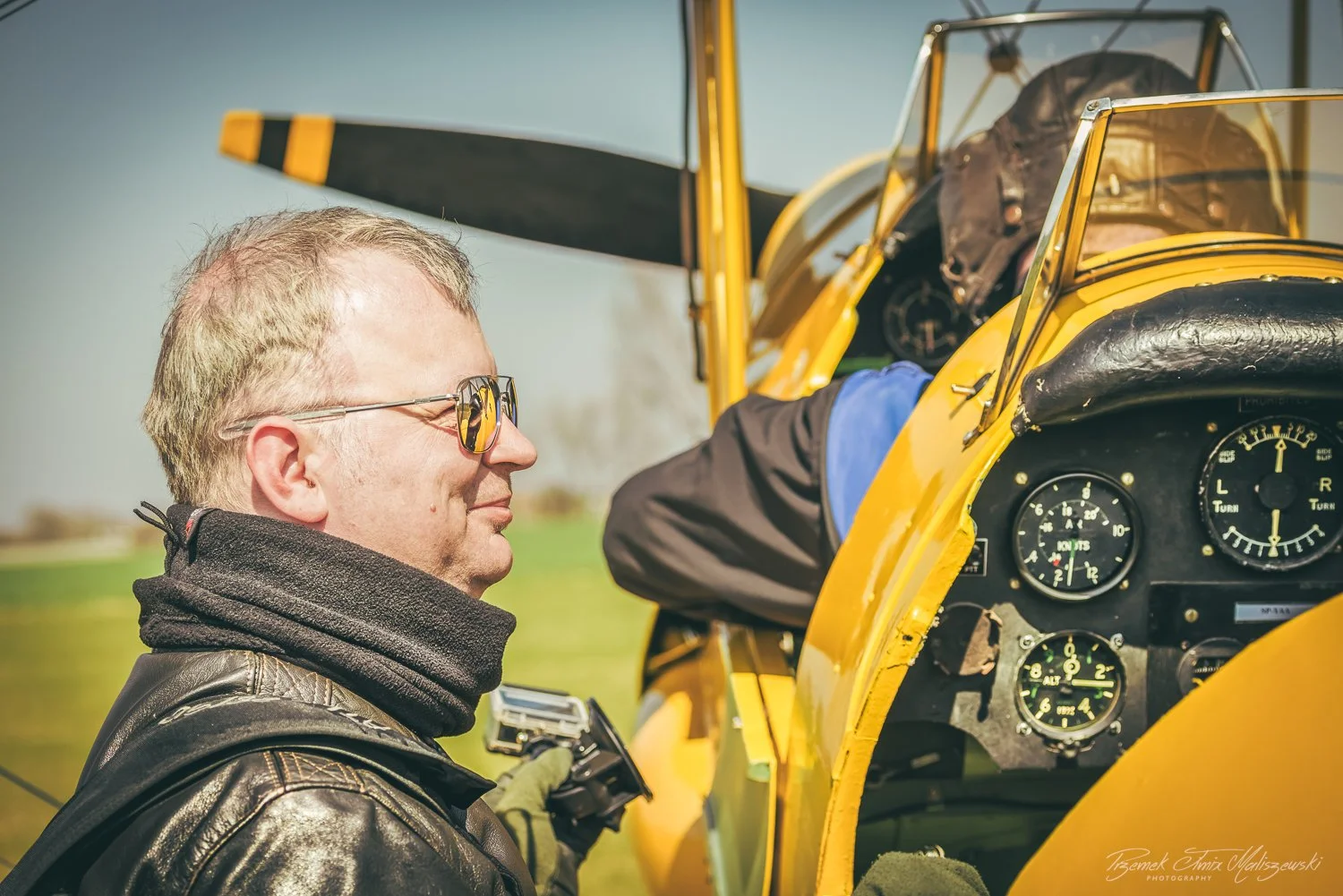 A man wearing sunglasses and a leather jacket standing next to a yellow vintage airplane, holding a walkie-talkie, while another person is reaching into the aircraft's cockpit.