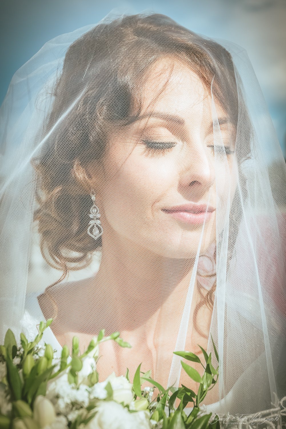 Close-up of a woman with closed eyes, wearing wedding earrings, covered partially by a veil, holding a bouquet of white flowers.