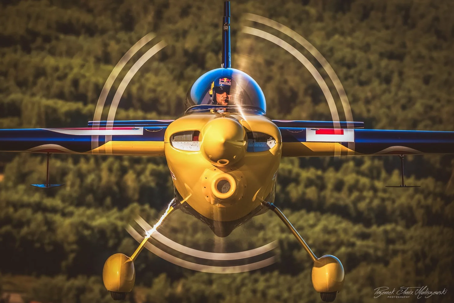 A yellow and black jet aircraft flying towards the camera, with a pilot wearing a helmet visible inside the cockpit. The aircraft's nose is prominent, and the background shows a blurred landscape of trees.