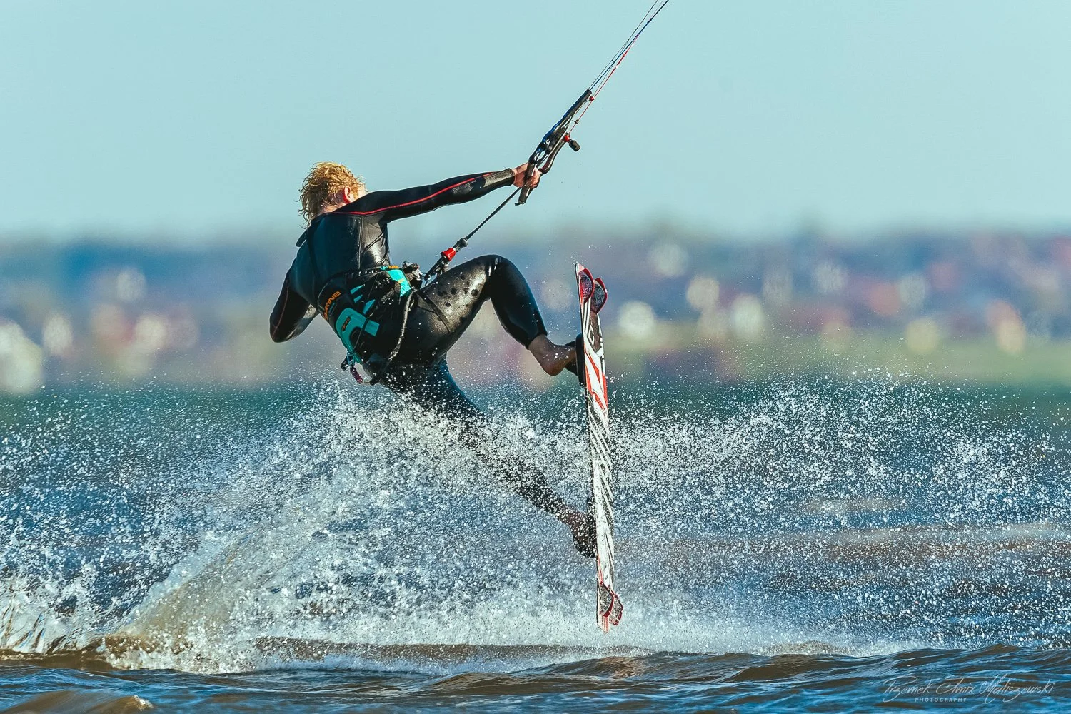 A person in a wetsuit performing a jump while kiteboarding over water, with a blurred distant shoreline in the background.