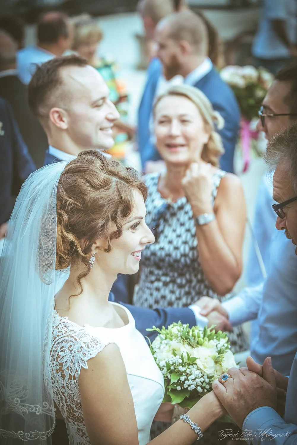 A bride and groom smiling and holding hands during a wedding ceremony surrounded by family and friends.