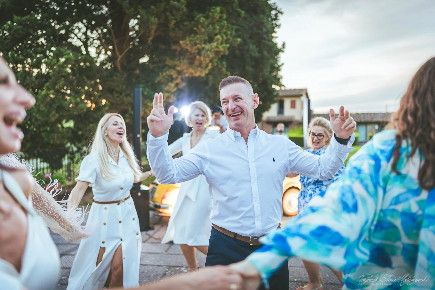 Group of people dancing and celebrating outdoors, smiling, with a man in a white shirt in the center, surrounded by women in various outfits, during a festive gathering in the evening.