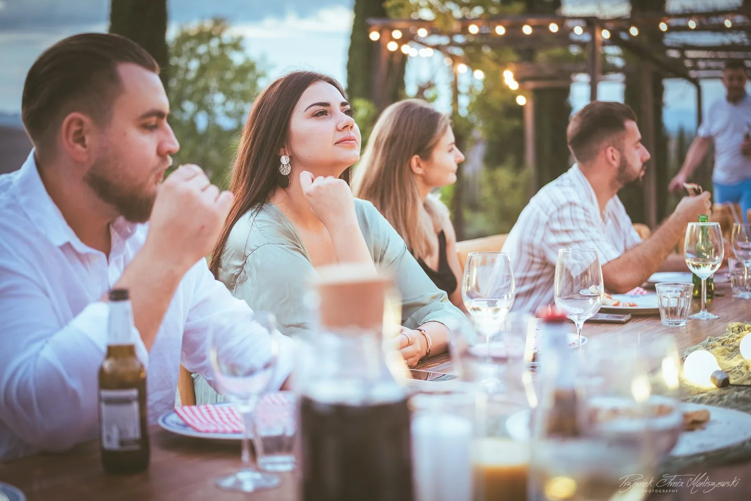 People sitting at a long outdoor dinner table in the evening, with various drinks and food, under string lights and trees.