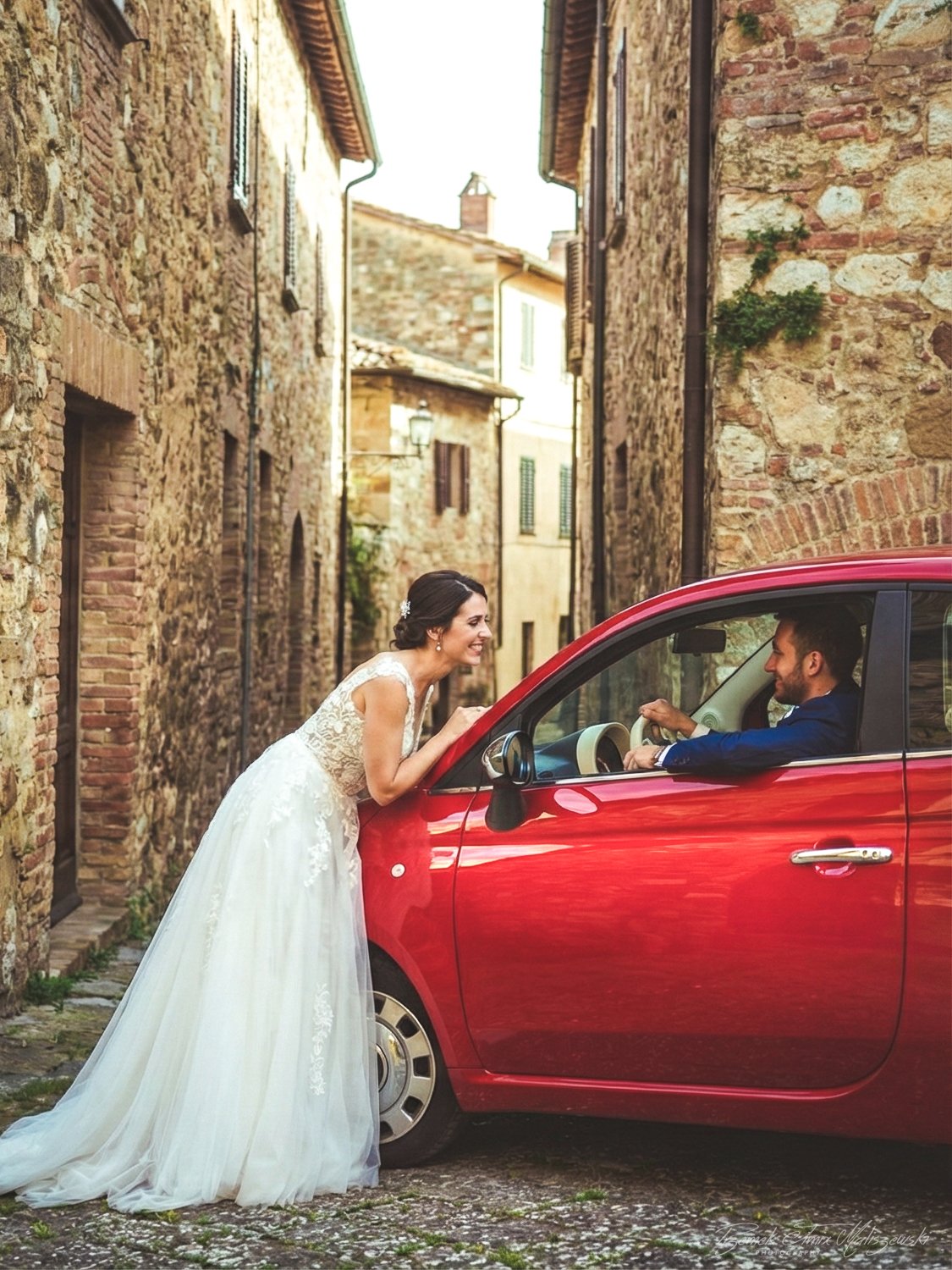 A bride in a white lace wedding dress leaning on a red car talking to a groom inside the car, in a narrow brick alley.