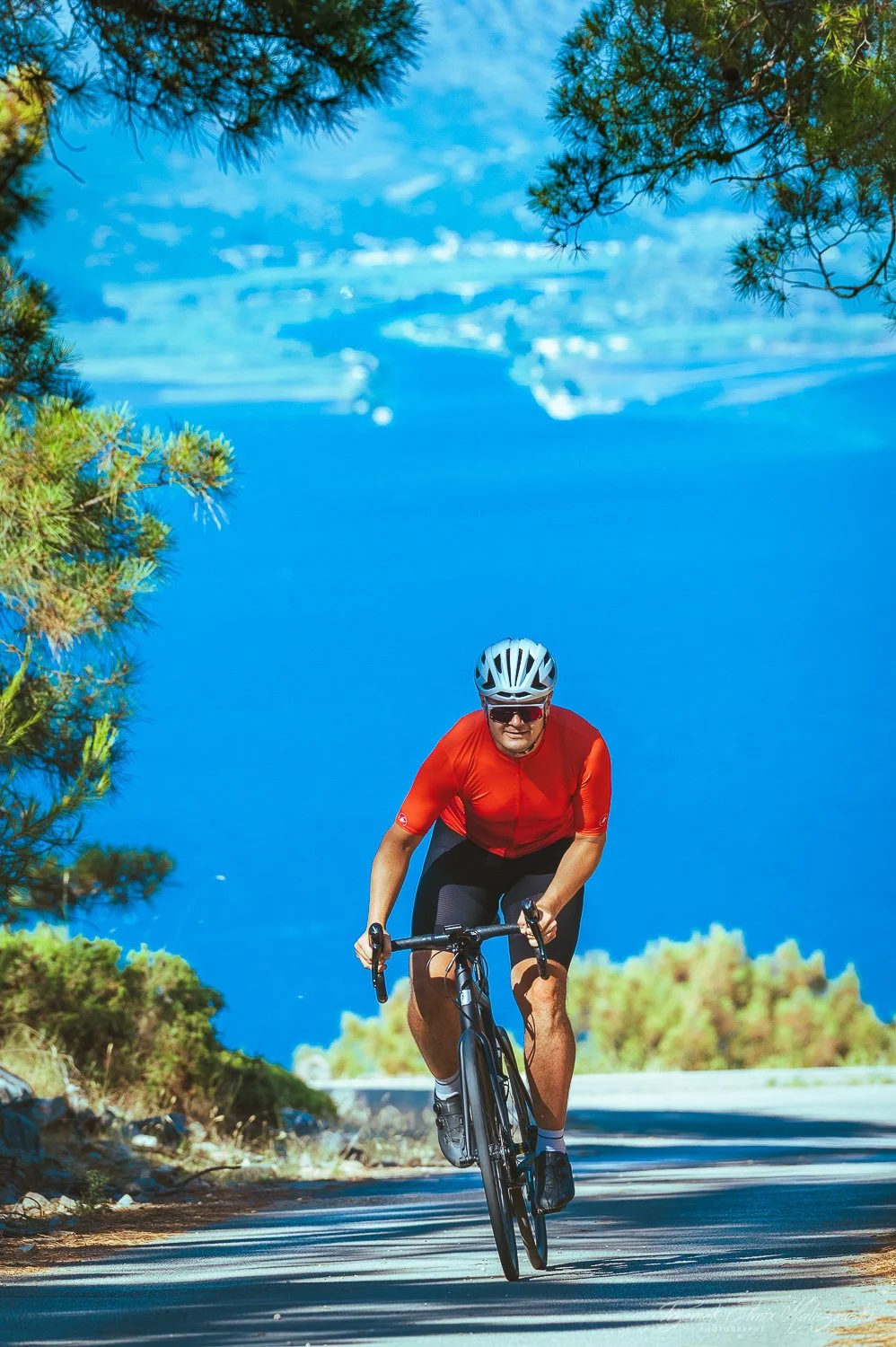 A man riding a black bicycle on a paved mountain road, wearing a red cycling jersey, black shorts, a white helmet, and sunglasses, with green trees in the foreground and a large body of water in the background.