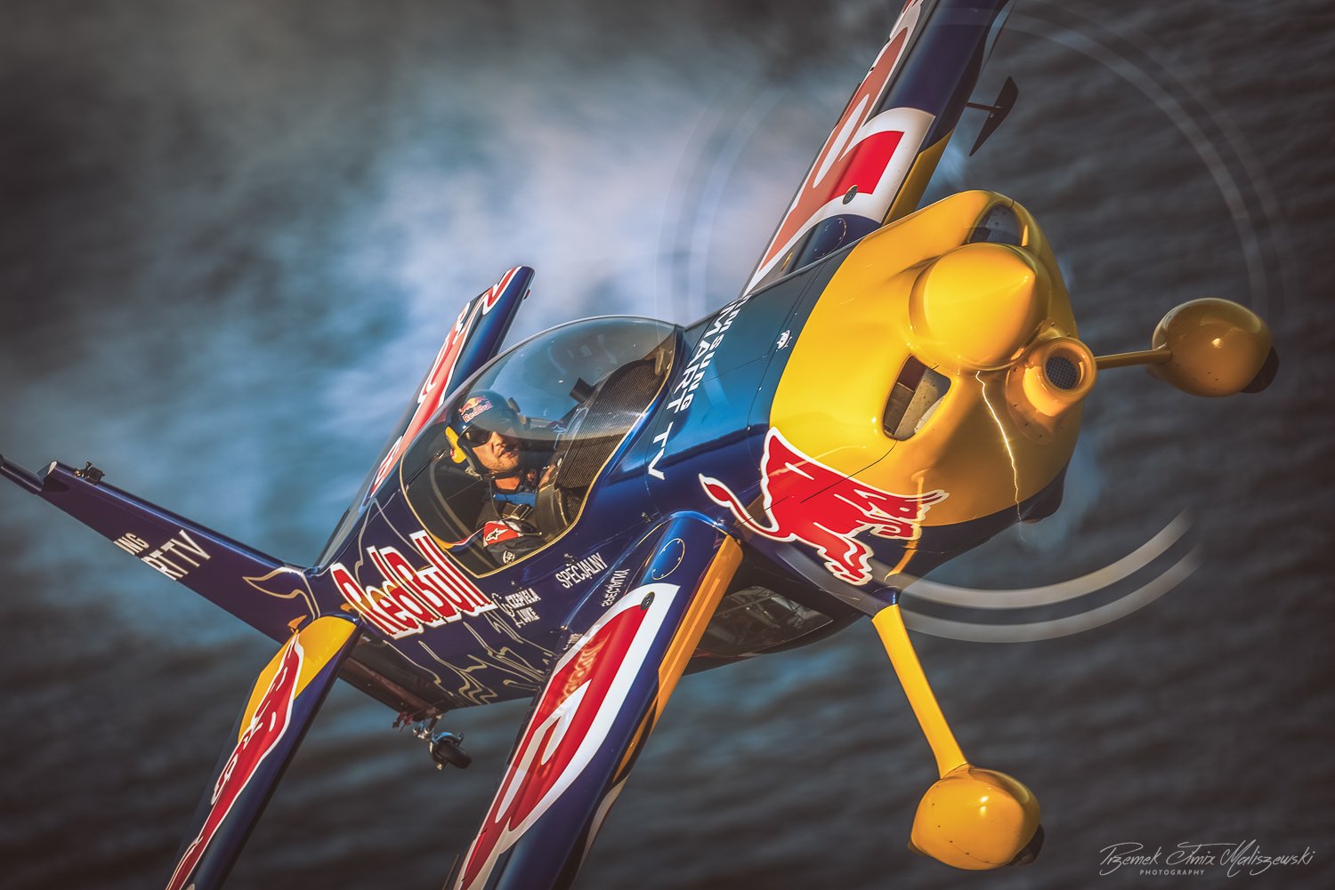 Close-up of a colorful aerobatic airplane with Red Bull branding in flight over water, with the pilot visible through the cockpit.