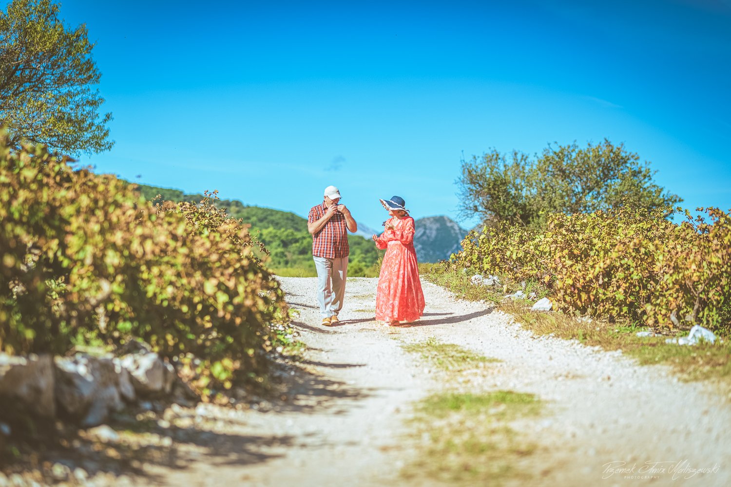 Two people, a man and a woman, walking on a dirt path outdoors on a sunny day, surrounded by bushes and green hills.