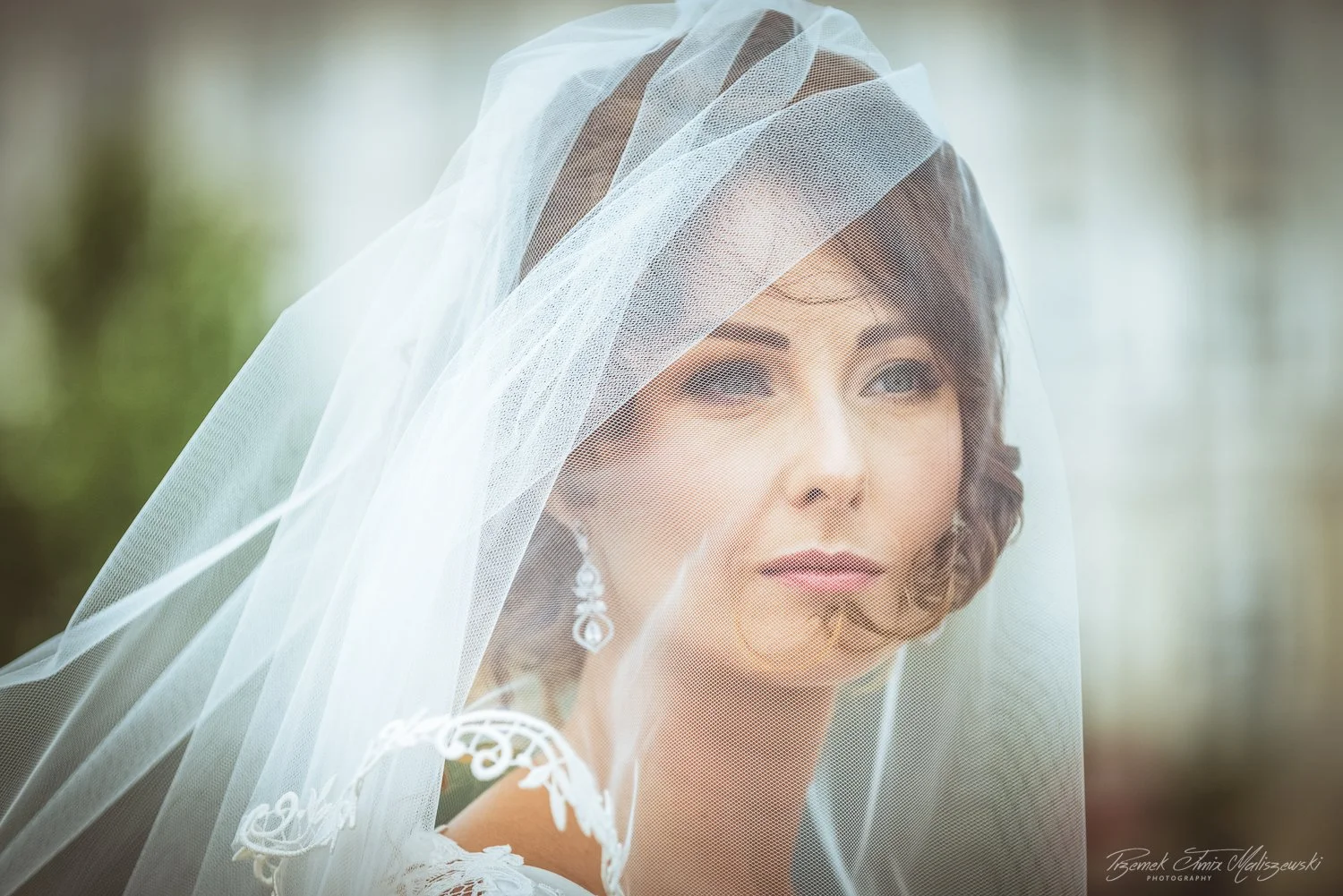A bride with short brown hair, wearing diamond earrings, looks thoughtfully into the distance with a white veil covering her face and shoulders.