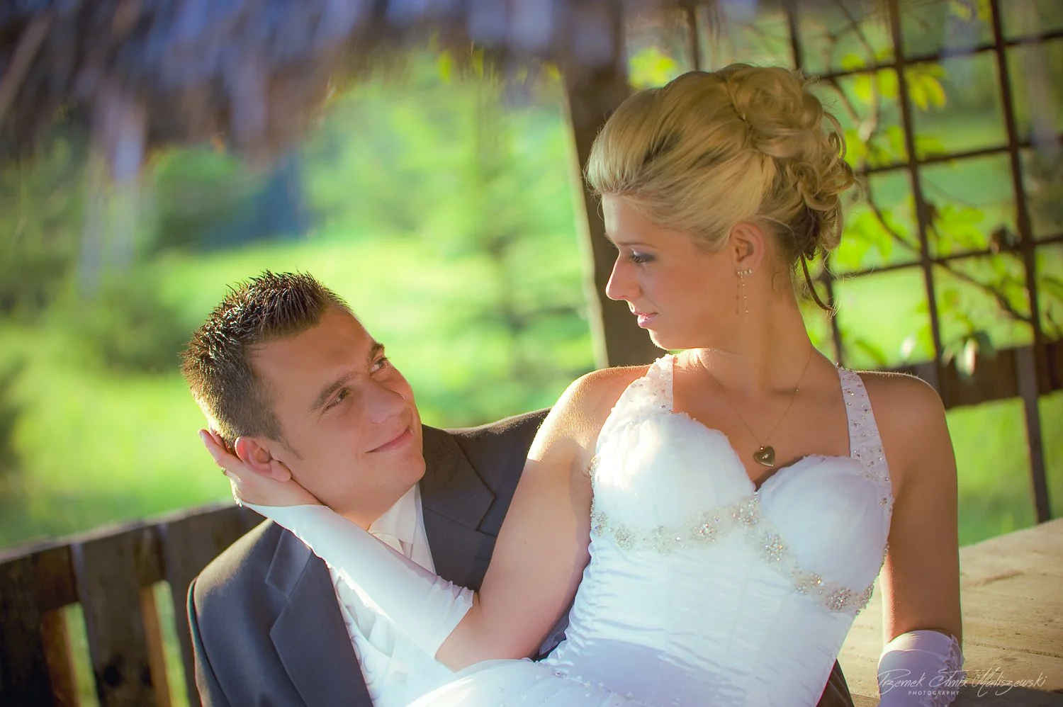 A newlywed couple sitting closely outdoors with a green, blurred background, the groom looking up at the bride who is gazing back at him.