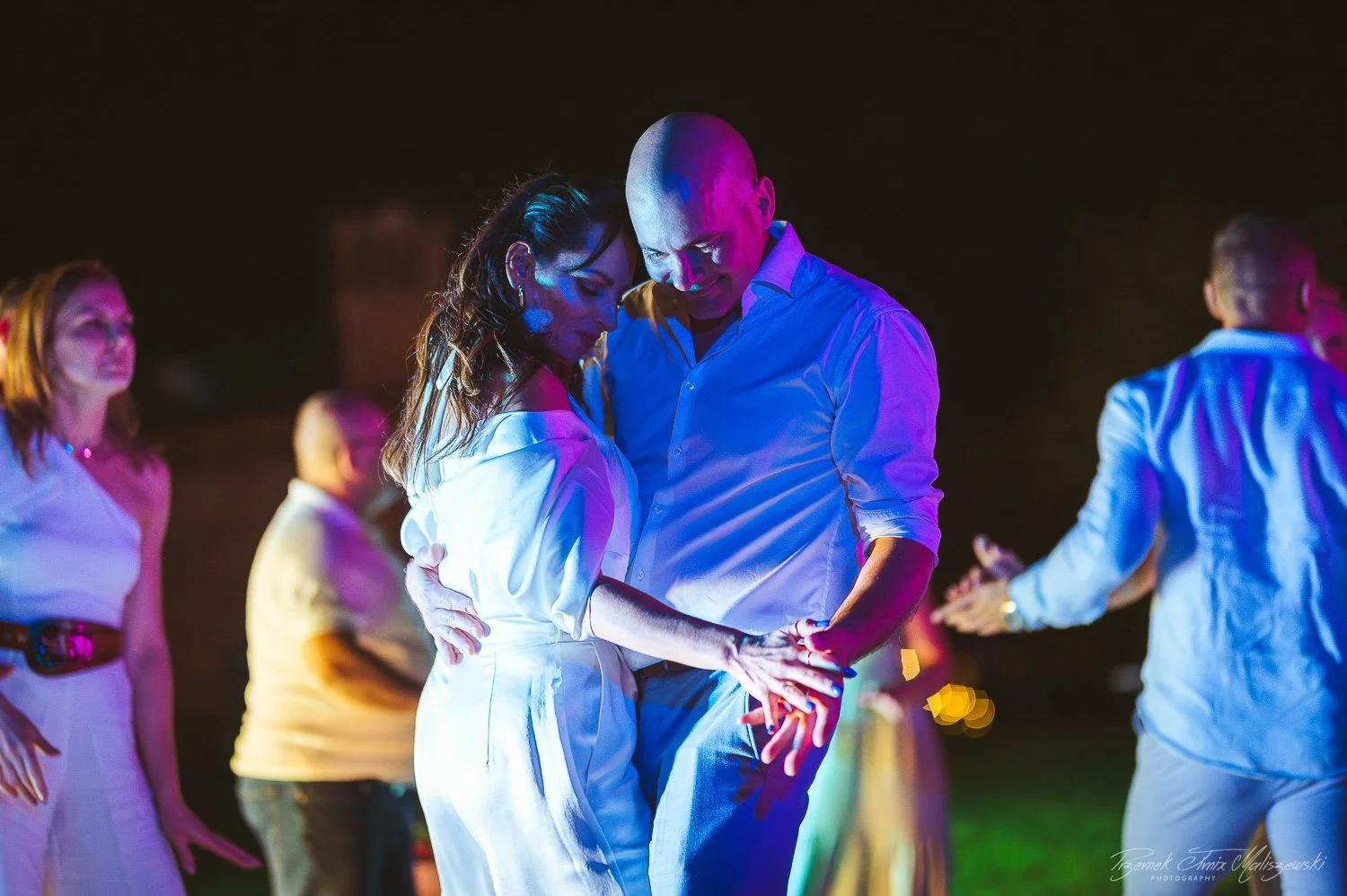 A man and woman dancing closely at an outdoor evening event, illuminated by colorful lights, with other people dancing in the background.