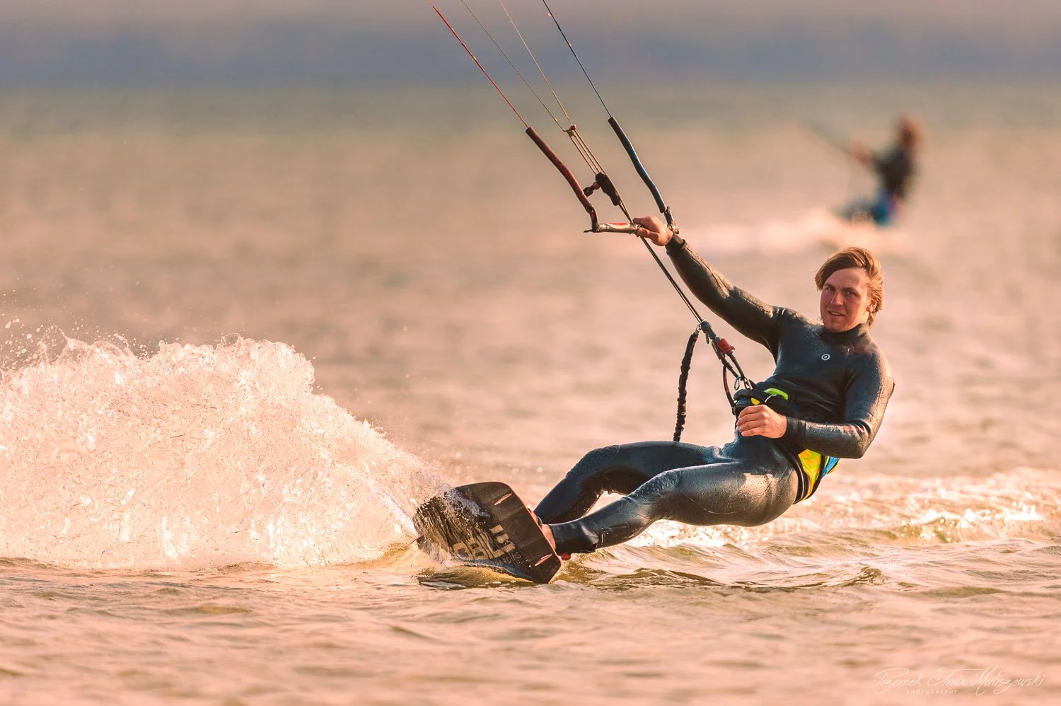 A man in a wetsuit kite surfing on the water during sunset, with another kite surfer in the background.