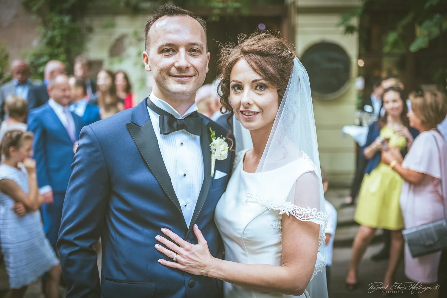 A bride and groom posing for a photo at their wedding, with guests in the background.
