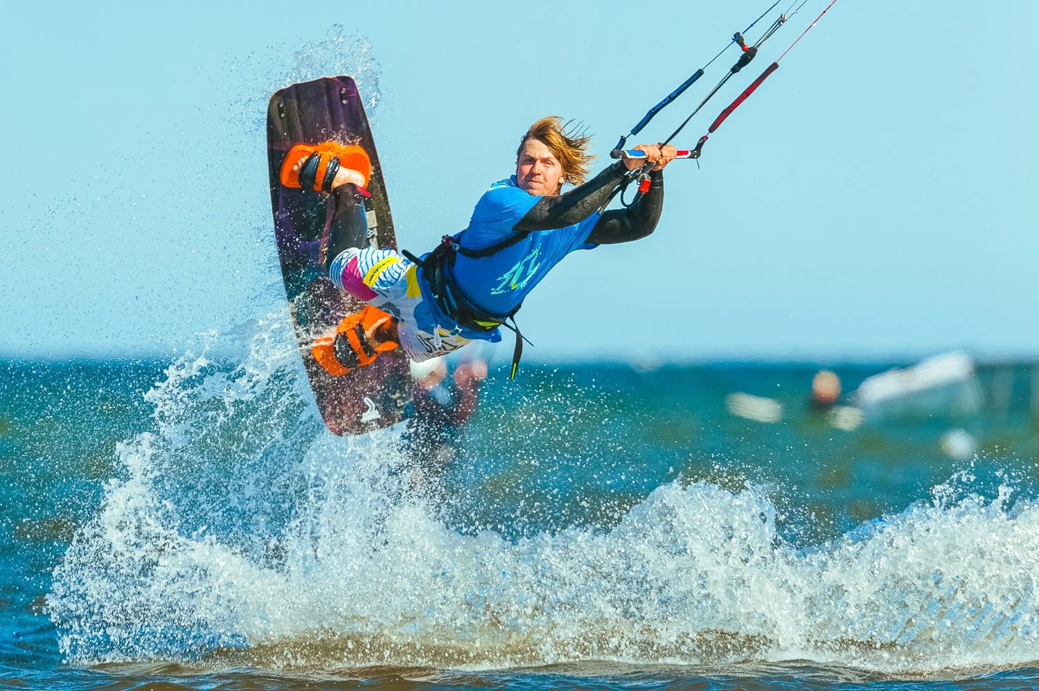 A man kiteboarding on the ocean, airborne above the water with a colorful kite and wearing a blue wetsuit and colorful shorts.