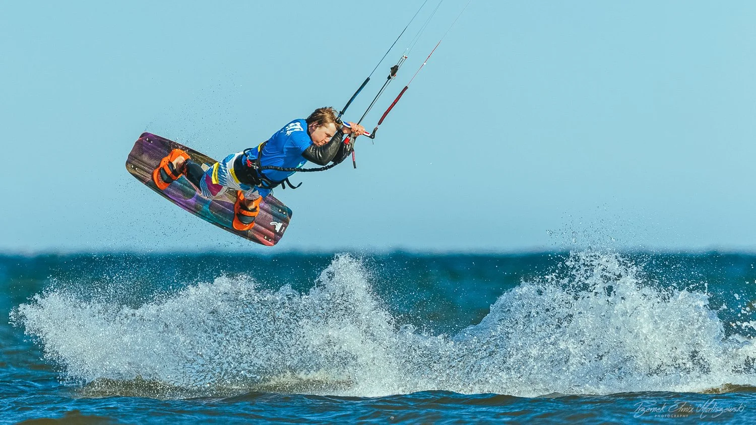 A person kiteboarding above the water, wearing a blue shirt and colorful shorts, holding the control bar, with water splashing below.