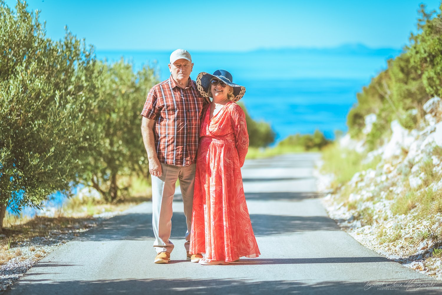 An elderly man and woman standing on a road near the water, dressed in summer clothing and smiling at the camera, with greenery and the ocean in the background.