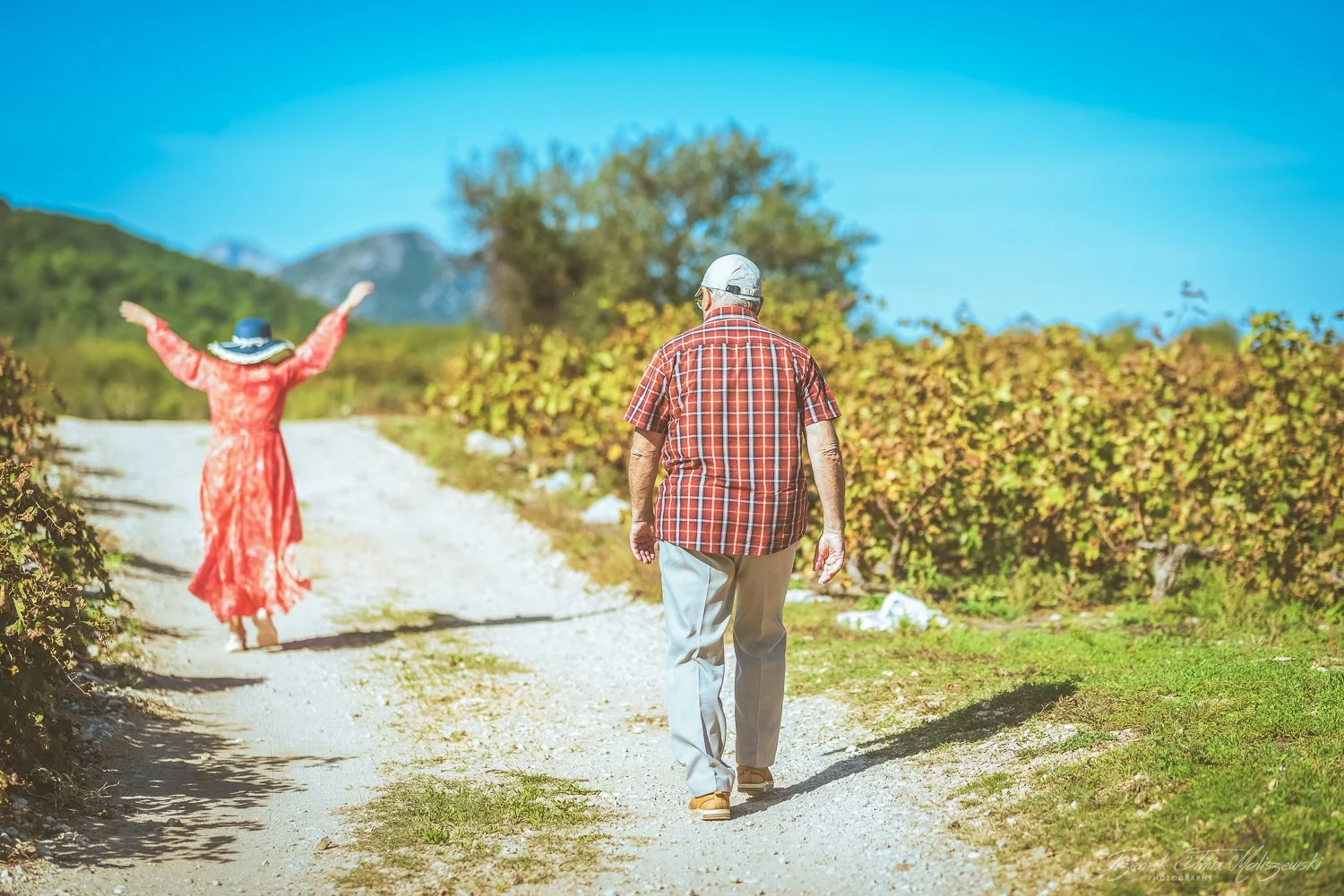 An elderly man in a plaid shirt and a woman in a red dress and large hat walking on a dirt path in a natural rural setting with bushes and trees, under a blue sky.