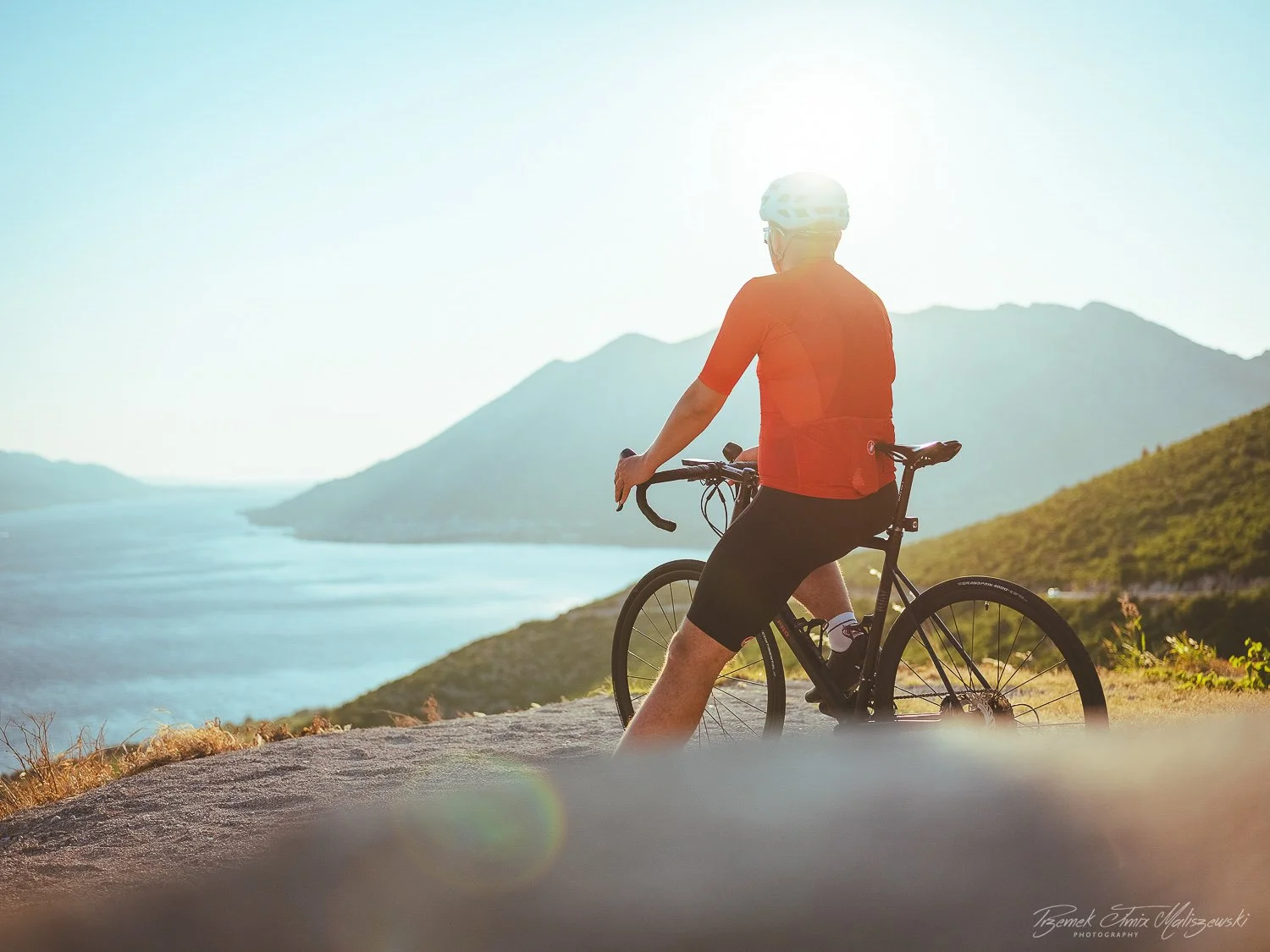 Man wearing a red shirt and black shorts riding a bicycle on a scenic mountain trail with water and mountains in the background during sunset.