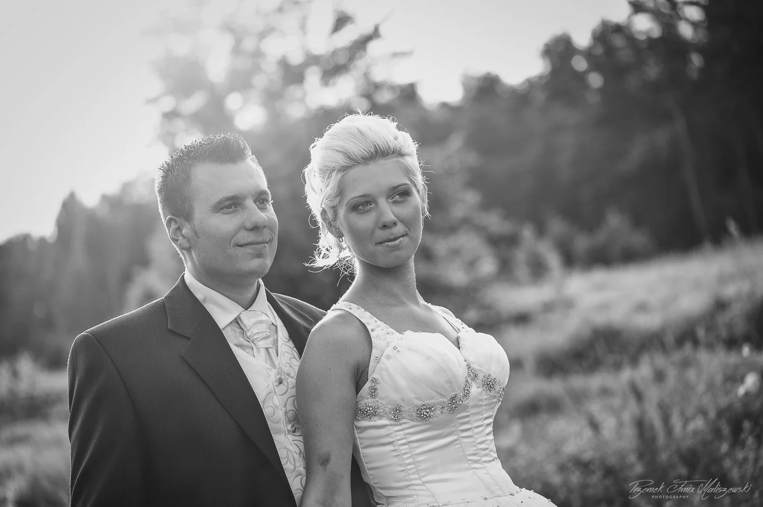 Black and white photo of a bride and groom outdoors. The groom in a suit looks fondly at the bride, who is in a wedding dress. Background shows trees and natural scenery.