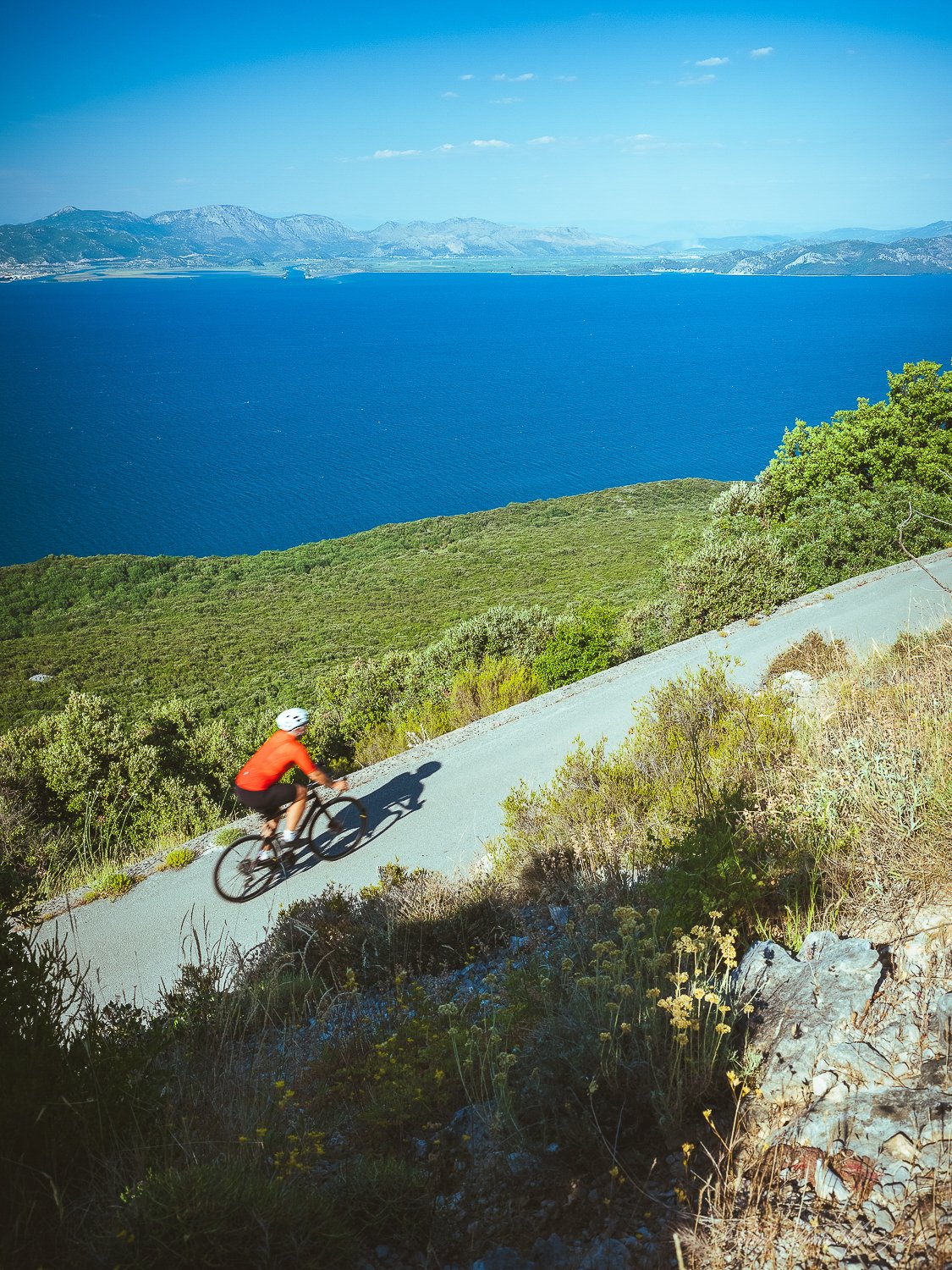 A cyclist wearing a red shirt and a white helmet riding a mountain bike on a steep, dirt trail overlooking a large blue lake with mountains in the background.