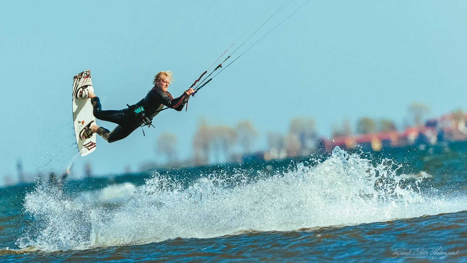 Person performing a jump on a wakeboard above the water surface during daytime with a distant shoreline in the background.