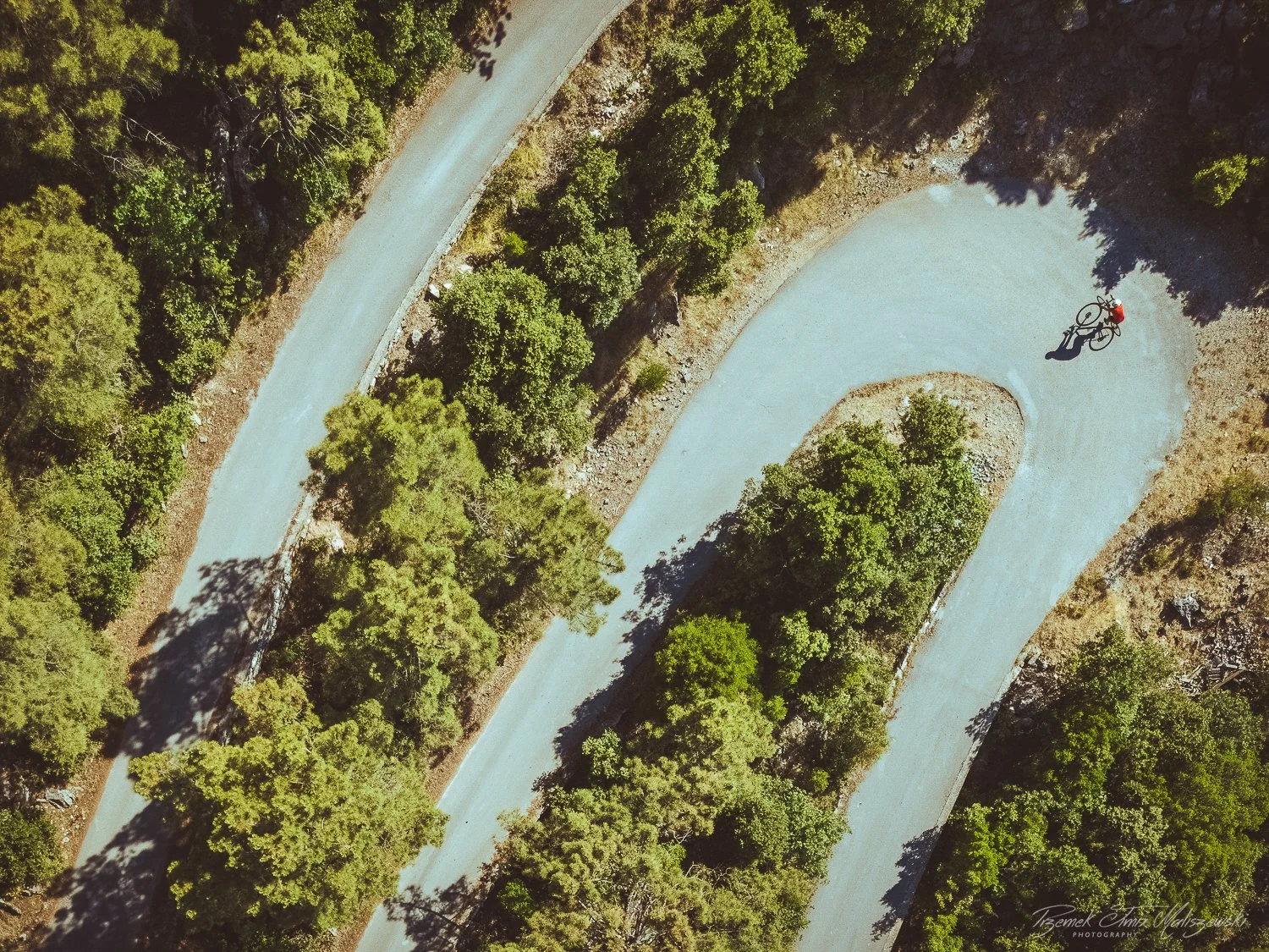 Aerial view of a winding dirt or gravel road surrounded by green trees with a single person riding a bicycle.