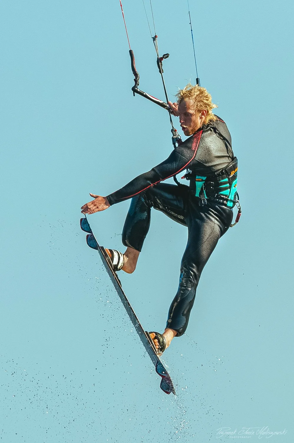A man engaged in kiteboarding, mid-air, wearing a wetsuit, with a kite control bar, and water droplets visible below him against a clear blue sky.