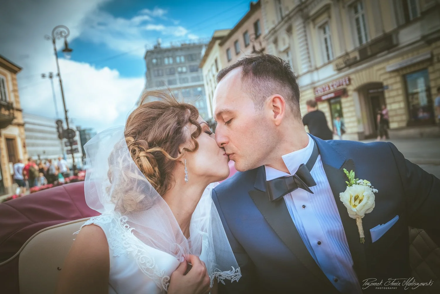 A bride and groom sharing a kiss during their wedding, sitting in a vintage car on a city street with historic buildings and pedestrians in the background.