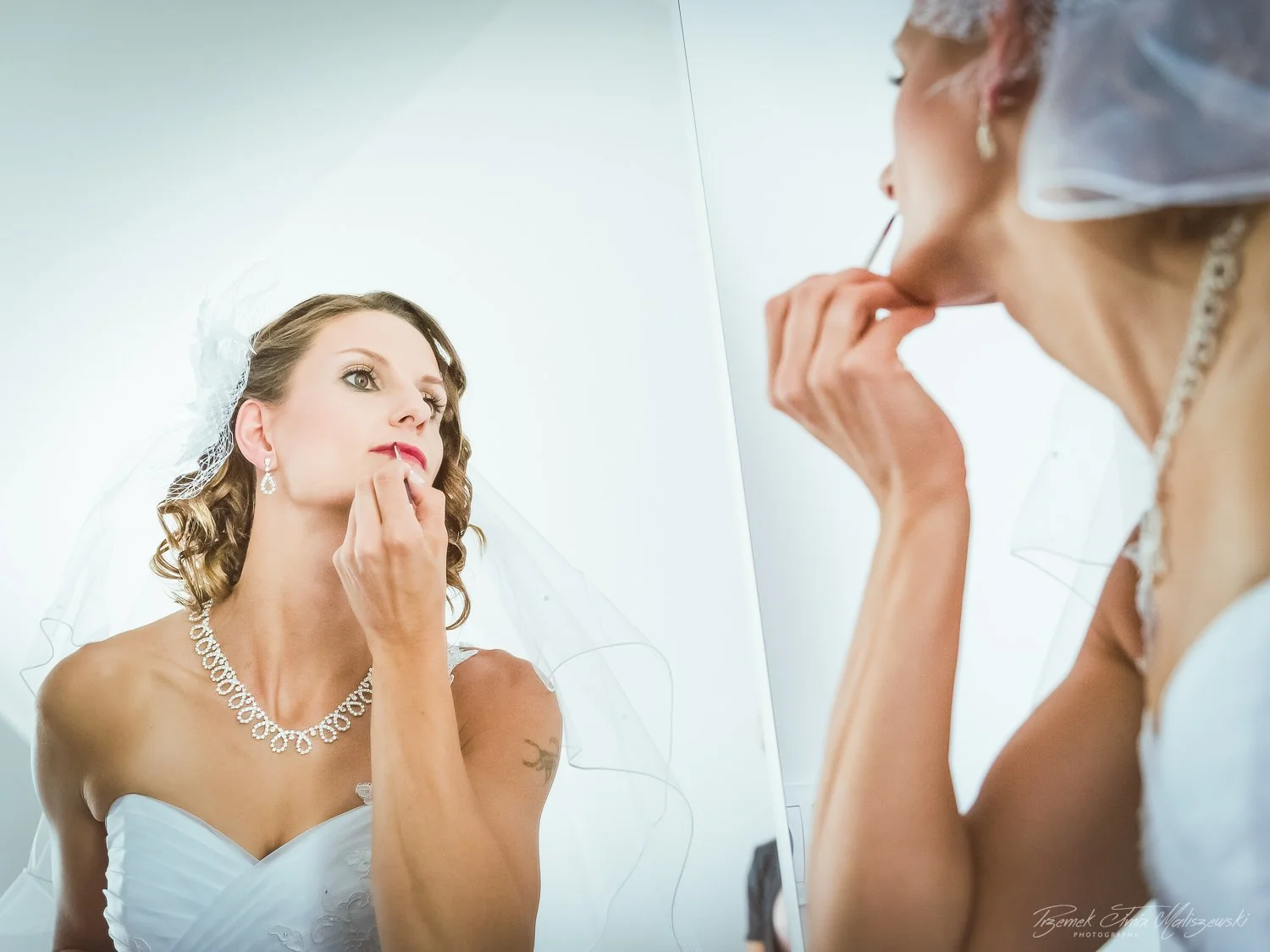 Bride with jewelry and veil applying lipstick in front of a mirror.