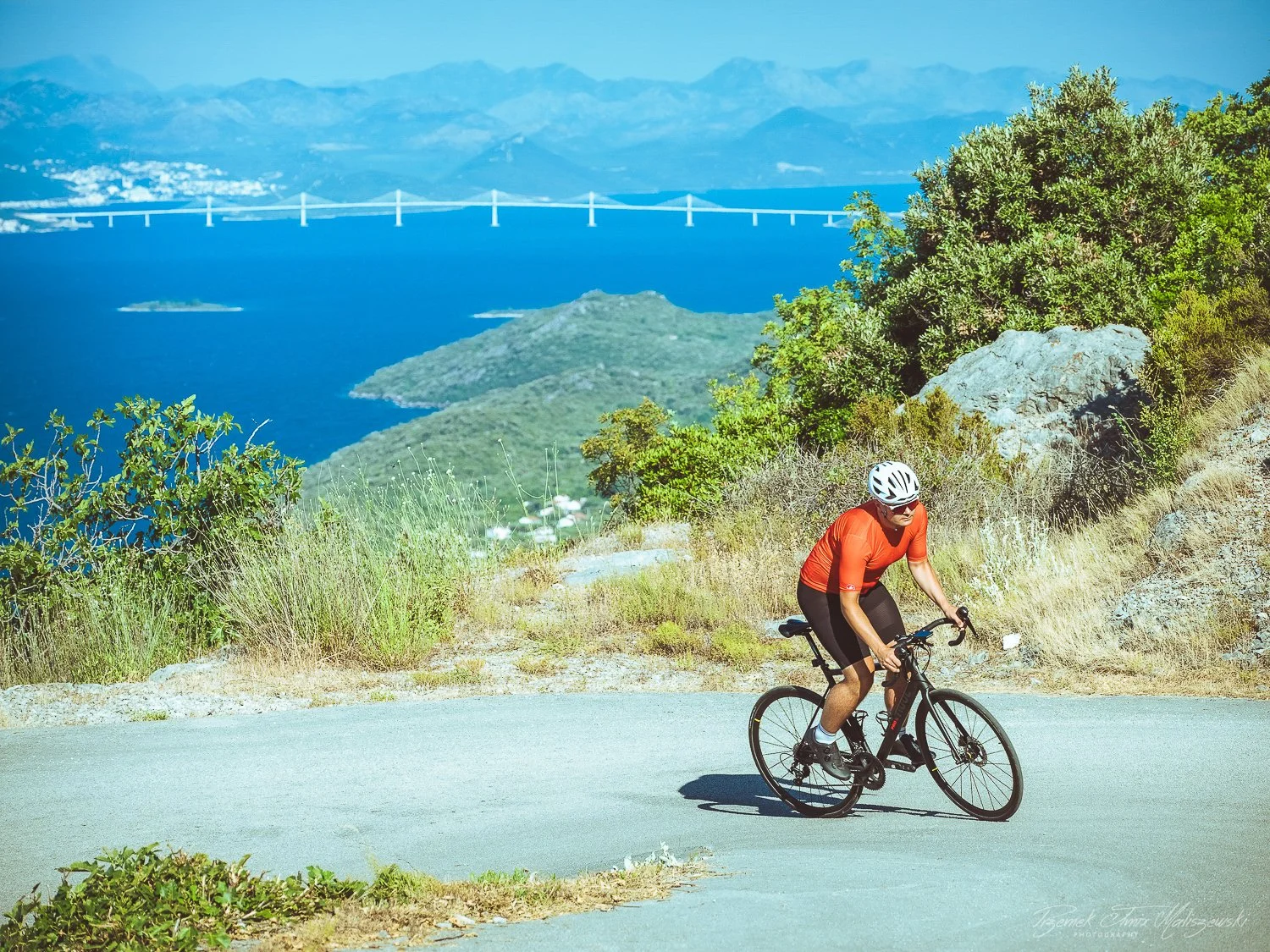 A man riding a bicycle on a mountain road with a scenic view of a blue lake, green hills, and a distant bridge and mountains in the background.