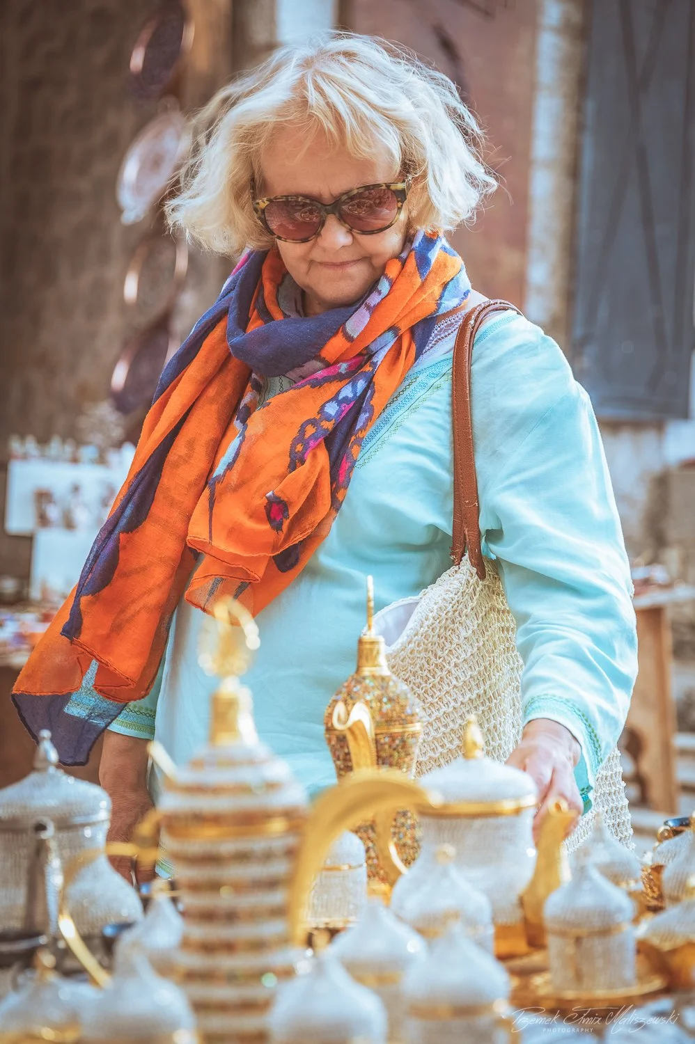 An older woman with curly blonde hair, wearing sunglasses, a light blue shirt, and an orange and blue patterned scarf, shopping at a market with various ornate teapots and containers.